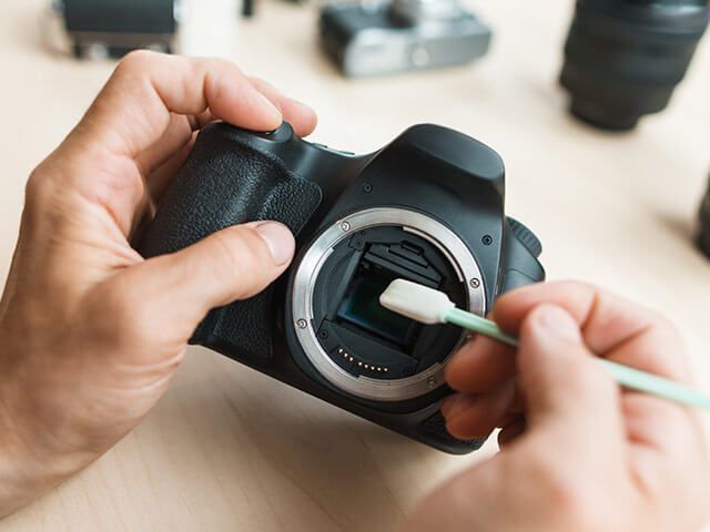 Des mains nettoient le capteur d'un appareil photo reflex numérique à l'aide d'un coton-tige. L'appareil est posé sur une surface en bois avec d'autres équipements photographiques.