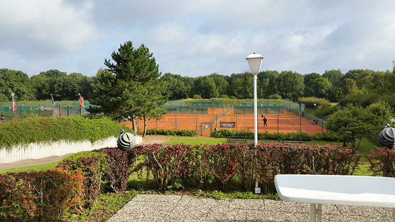 Ein Tennisplatz ist von Bäumen und Büschen umgeben und im Vordergrund steht ein Tisch.