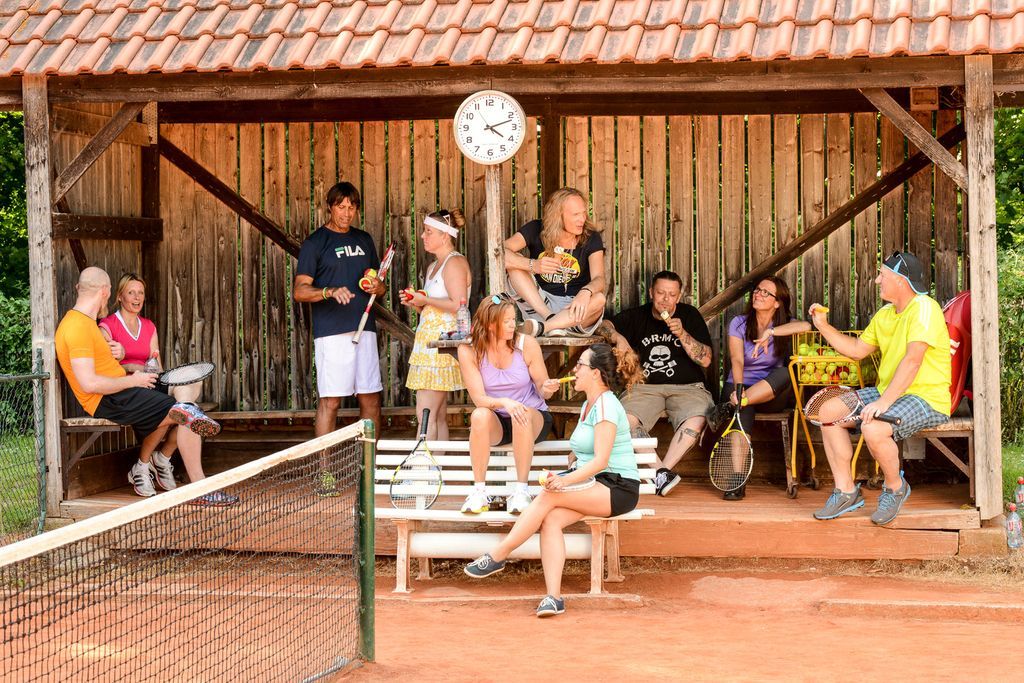Eine Gruppe von Menschen sitzt unter einem Holzunterstand auf einem Tennisplatz.