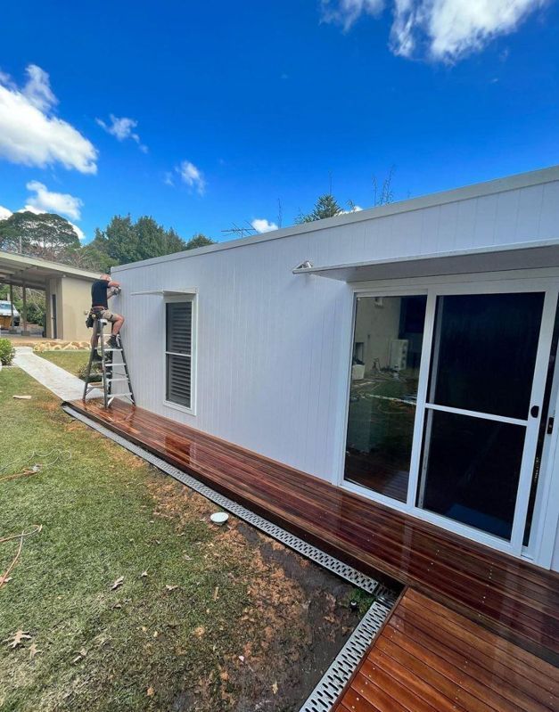 A man is standing on a ladder painting a window on a white house.