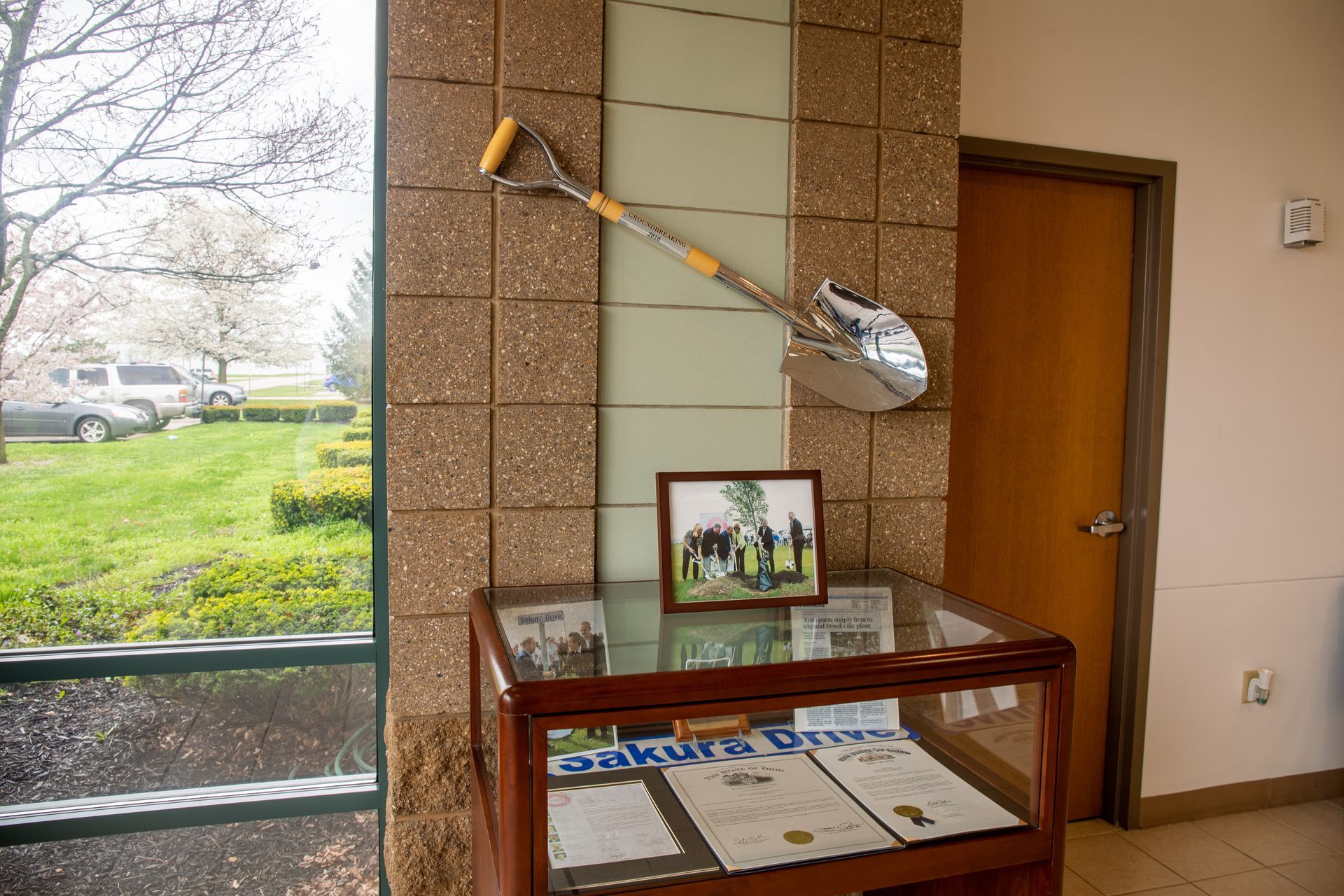 Shovel sculpture on wall, display case with documents, framed photo. Window view of parked cars and lawn.
