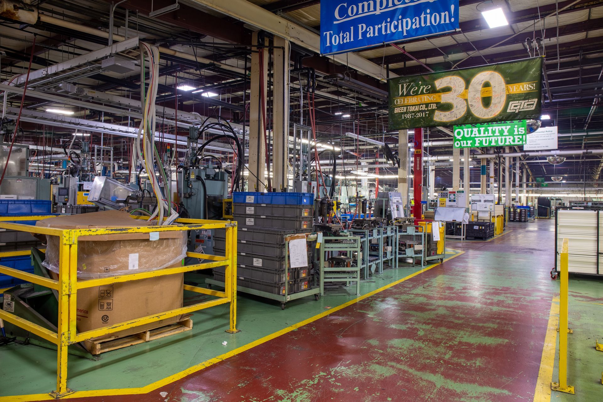 Factory interior with machinery, green floor, yellow safety rails, and signage.