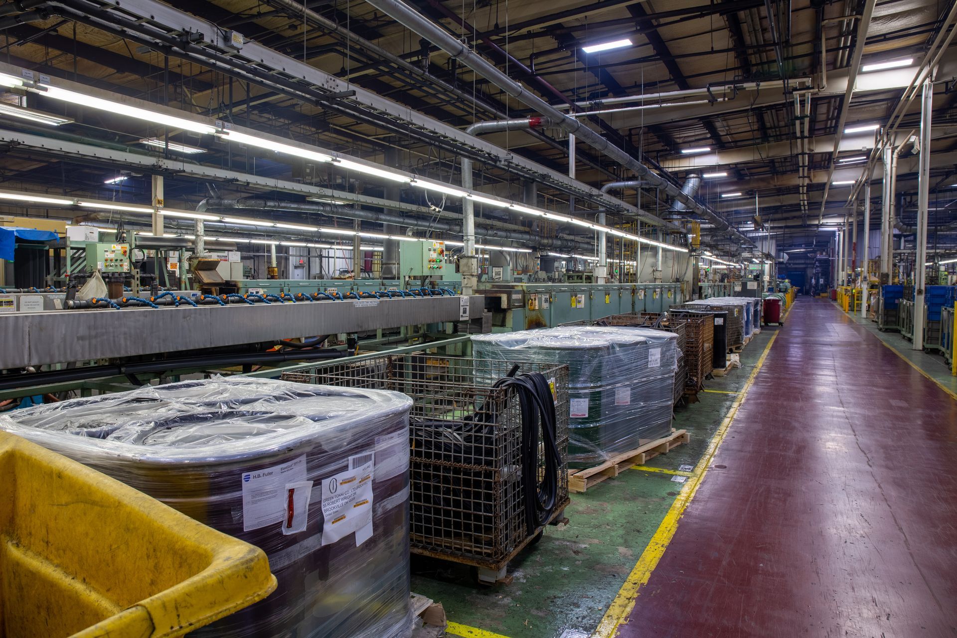 Factory interior with machinery, stacked packages, and a yellow lined aisle.