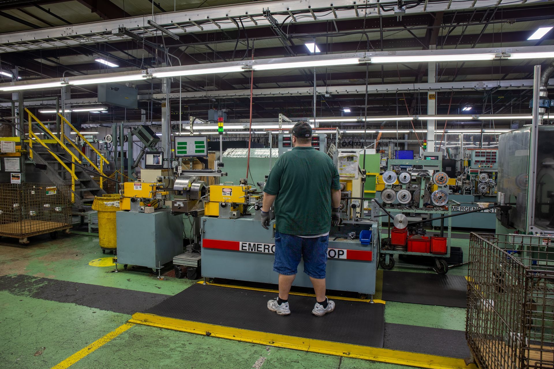 Man operating machinery in a factory. He stands on a mat facing a large machine with rolls of material.