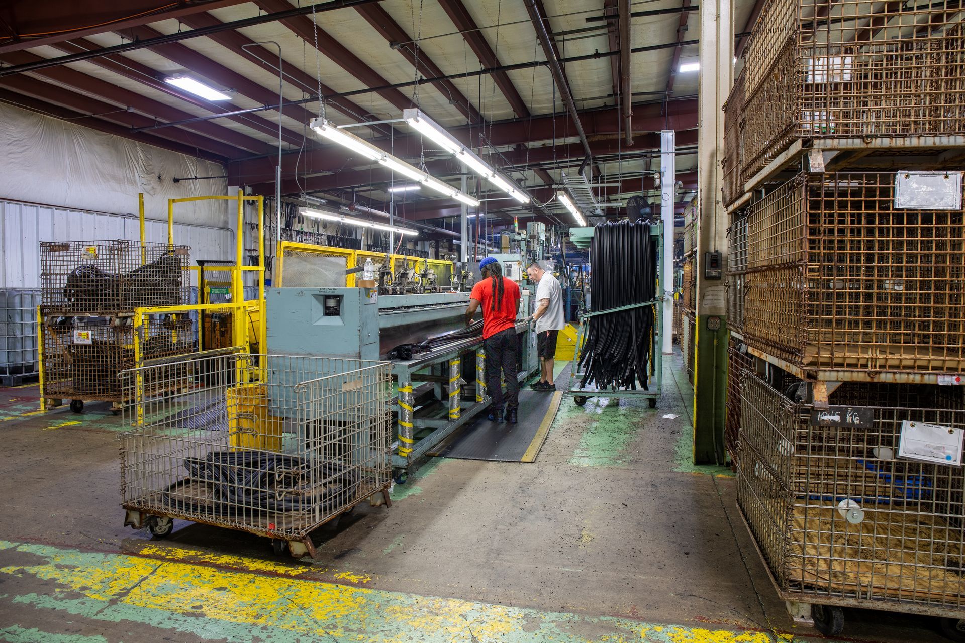 Factory interior with workers near machinery, metal cages stacked on pallets; fluorescent lighting.