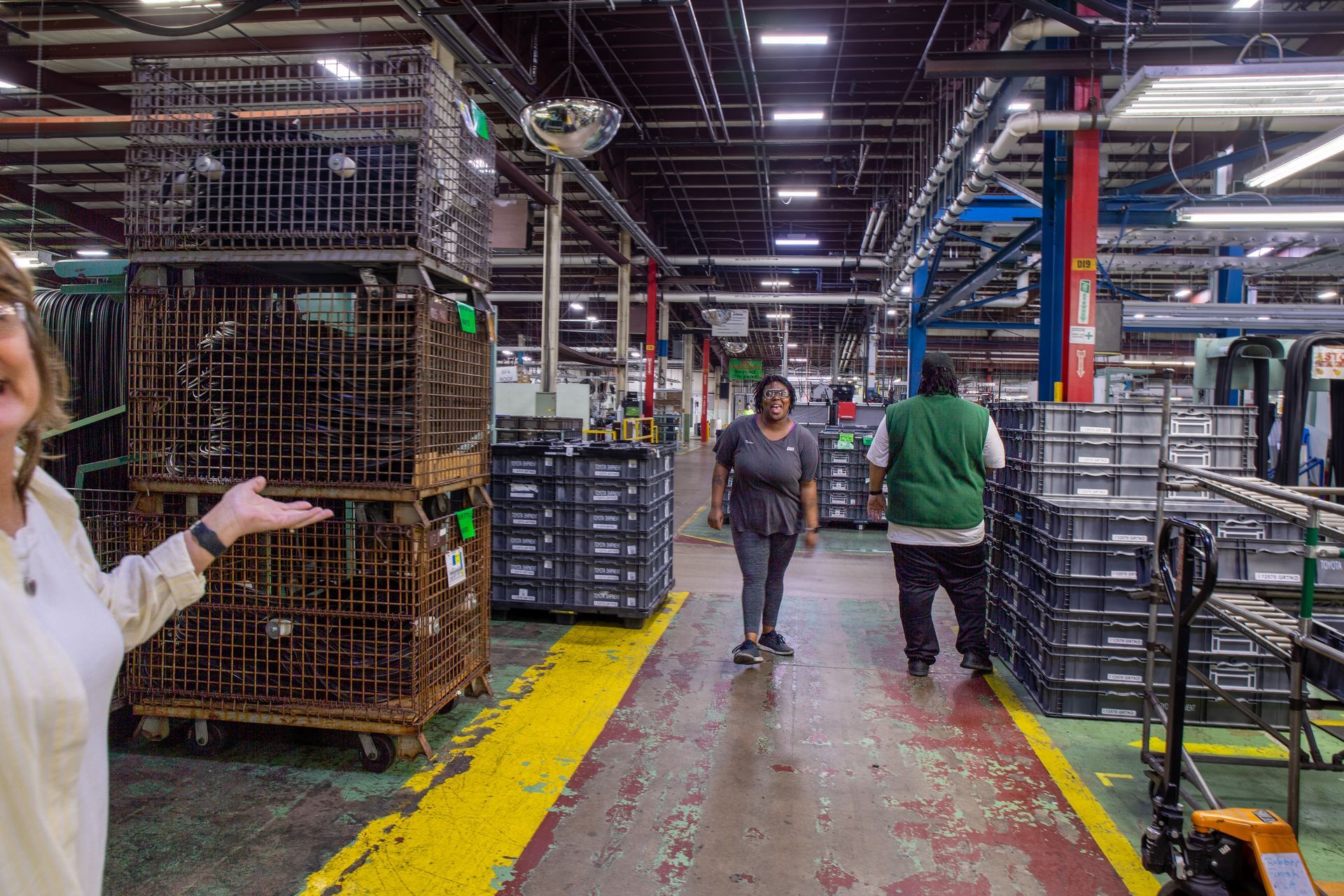 Factory interior with workers, stacked metal bins, and a woman gesturing.