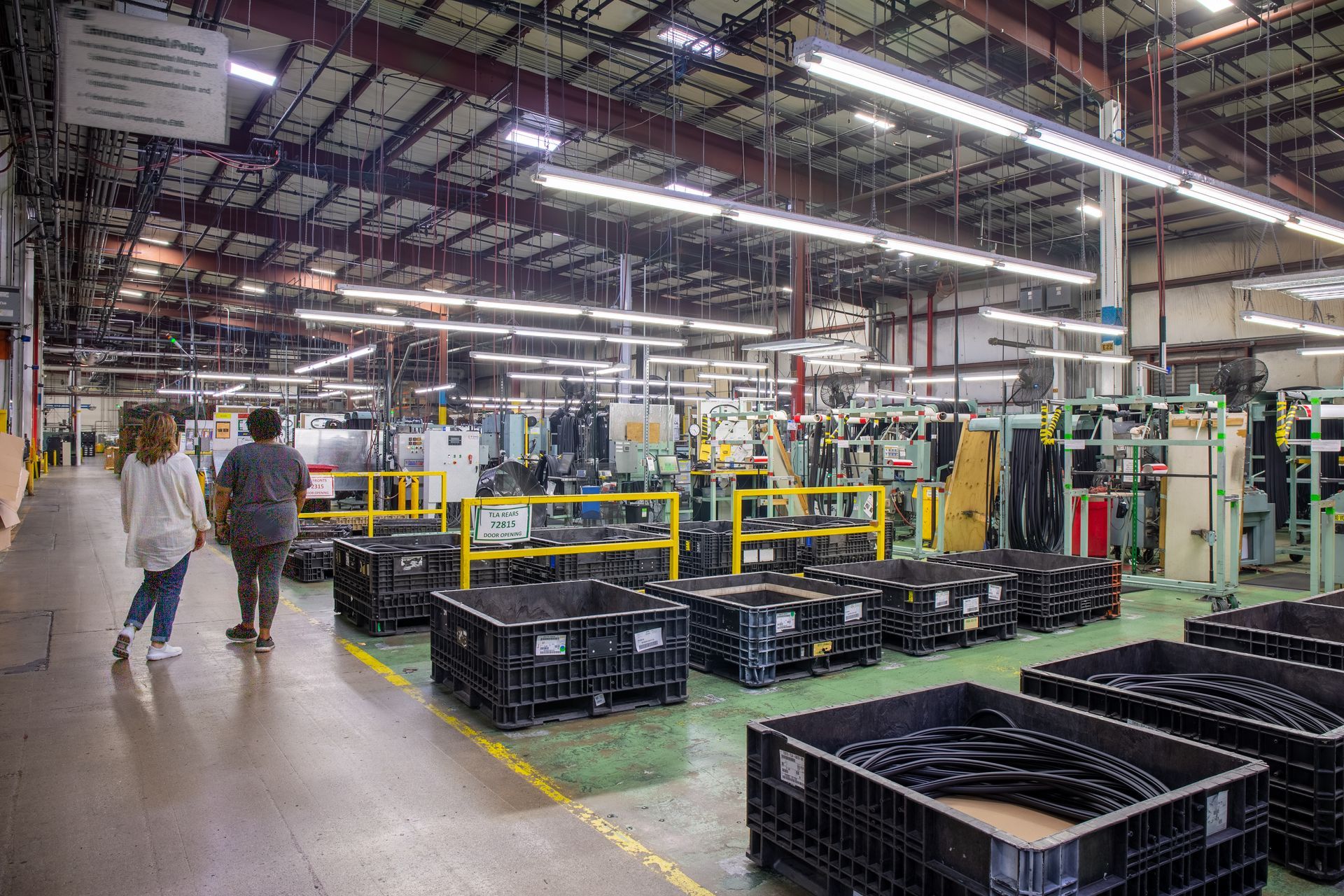 Two people walk through a factory, past machinery, and black containers on the floor.