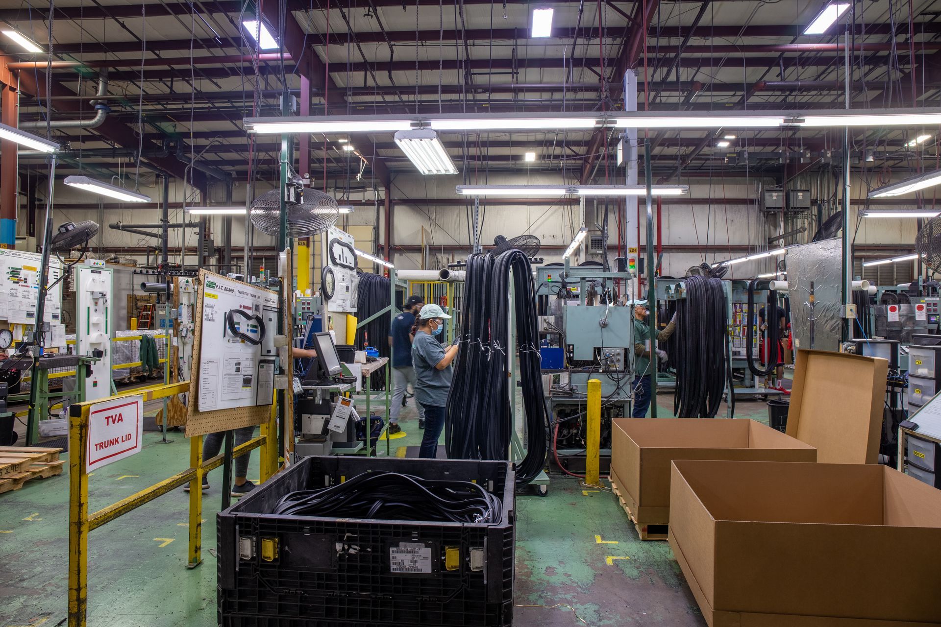 Factory interior with workers near machinery, assembly line, and cardboard boxes.