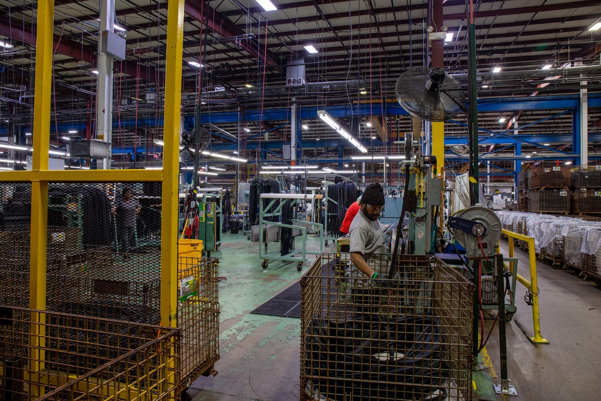 Factory interior with a worker at a workstation; yellow and blue machinery, metal baskets.