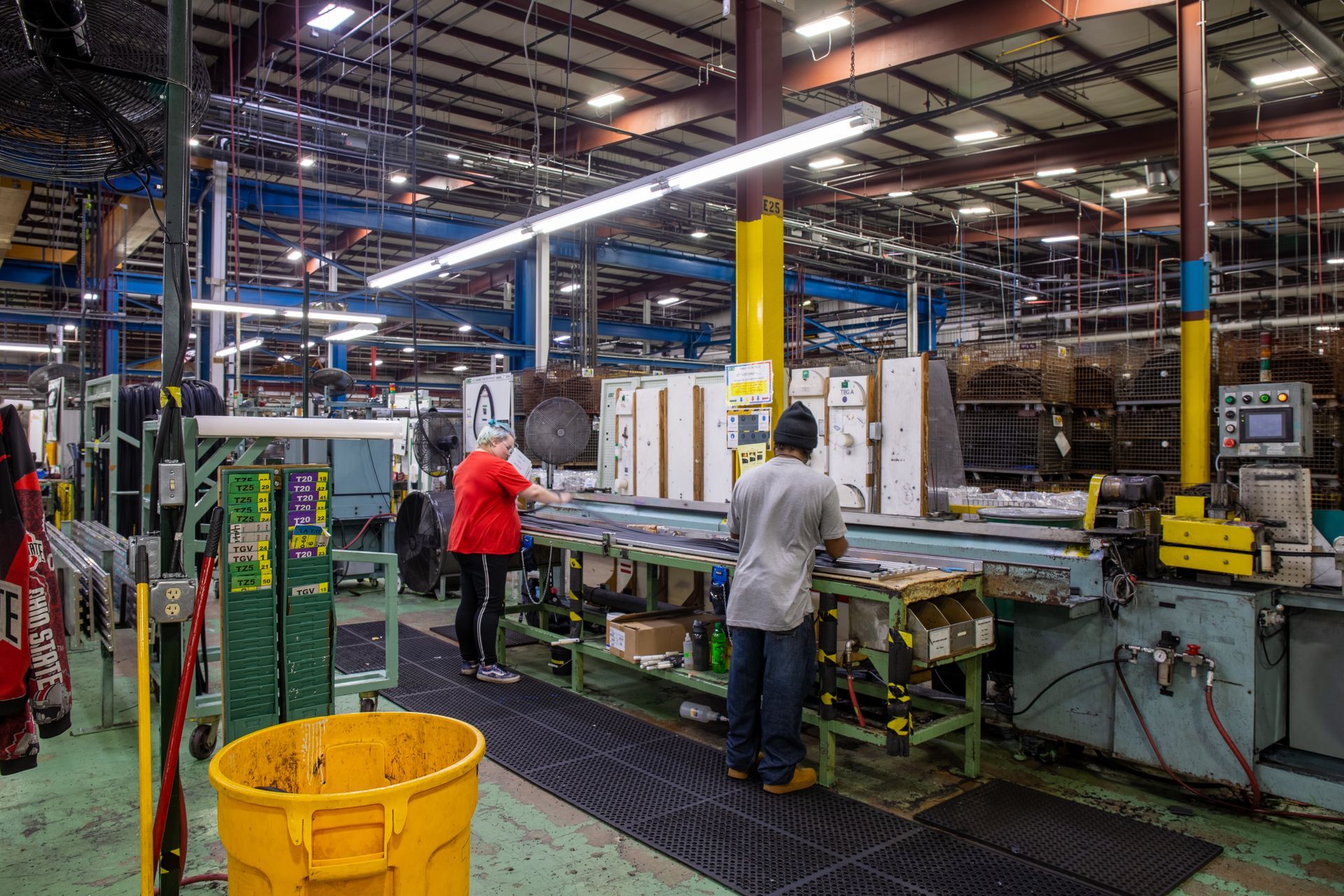 Factory interior with workers at a production line, with overhead lighting, a yellow trash can, and various machinery.
