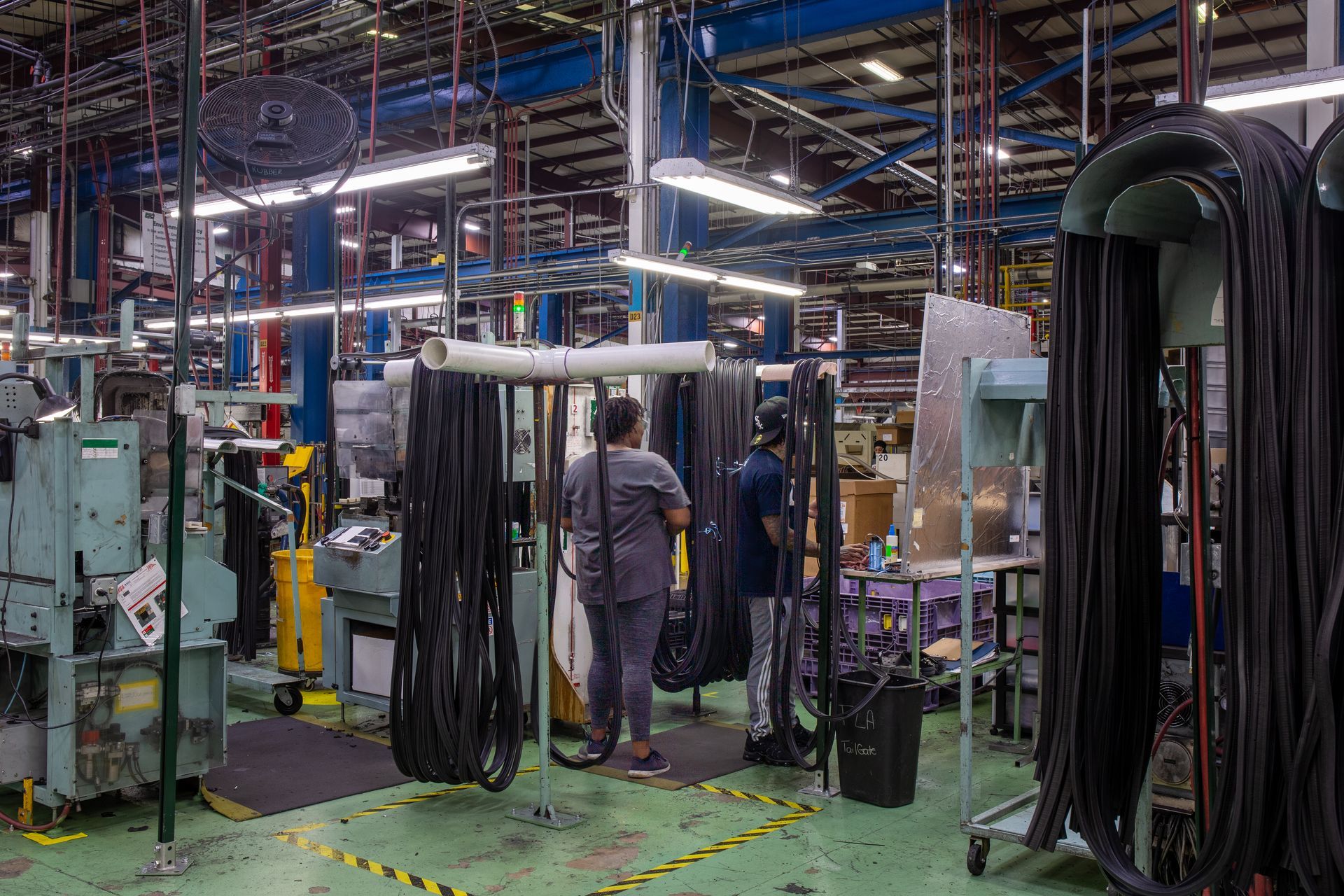 Two people standing in a factory. Black rubber tubes hang from racks. Machinery and equipment in background.