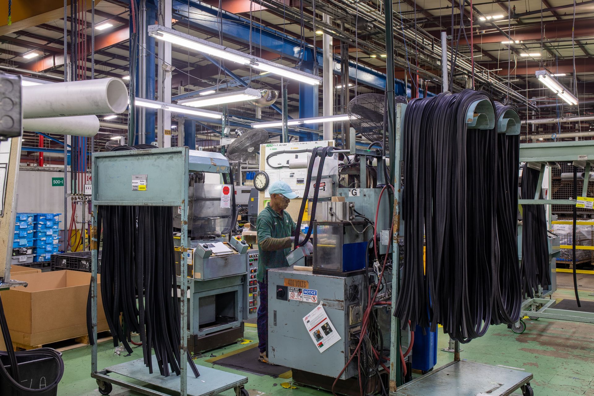Factory worker operating machinery with hanging black components. Industrial setting with overhead pipes.