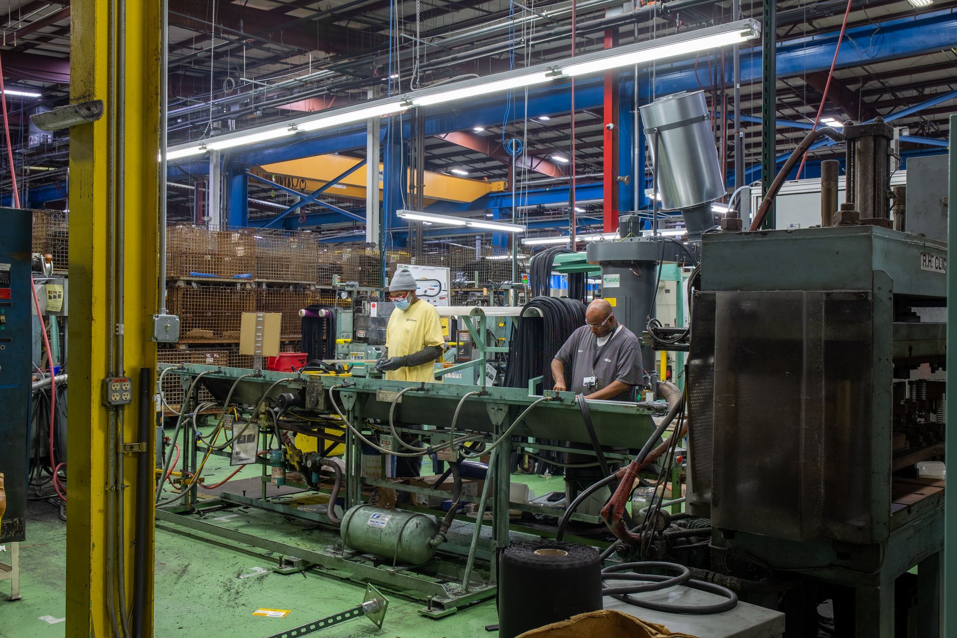 Two workers operate machinery in a factory. Green and gray equipment, bright overhead lights.