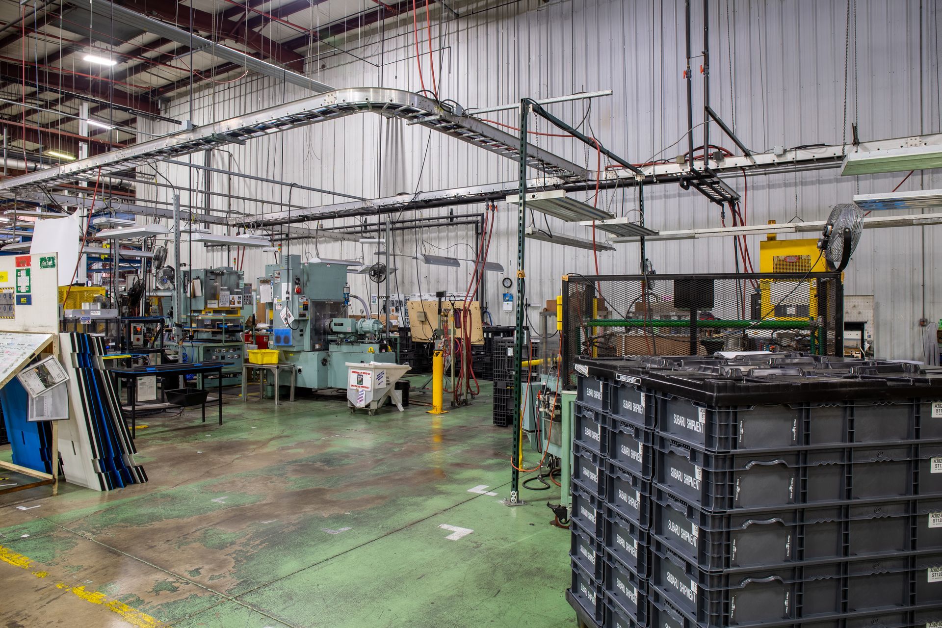 Industrial factory floor with machinery, overhead conveyor system, and stacked black bins.