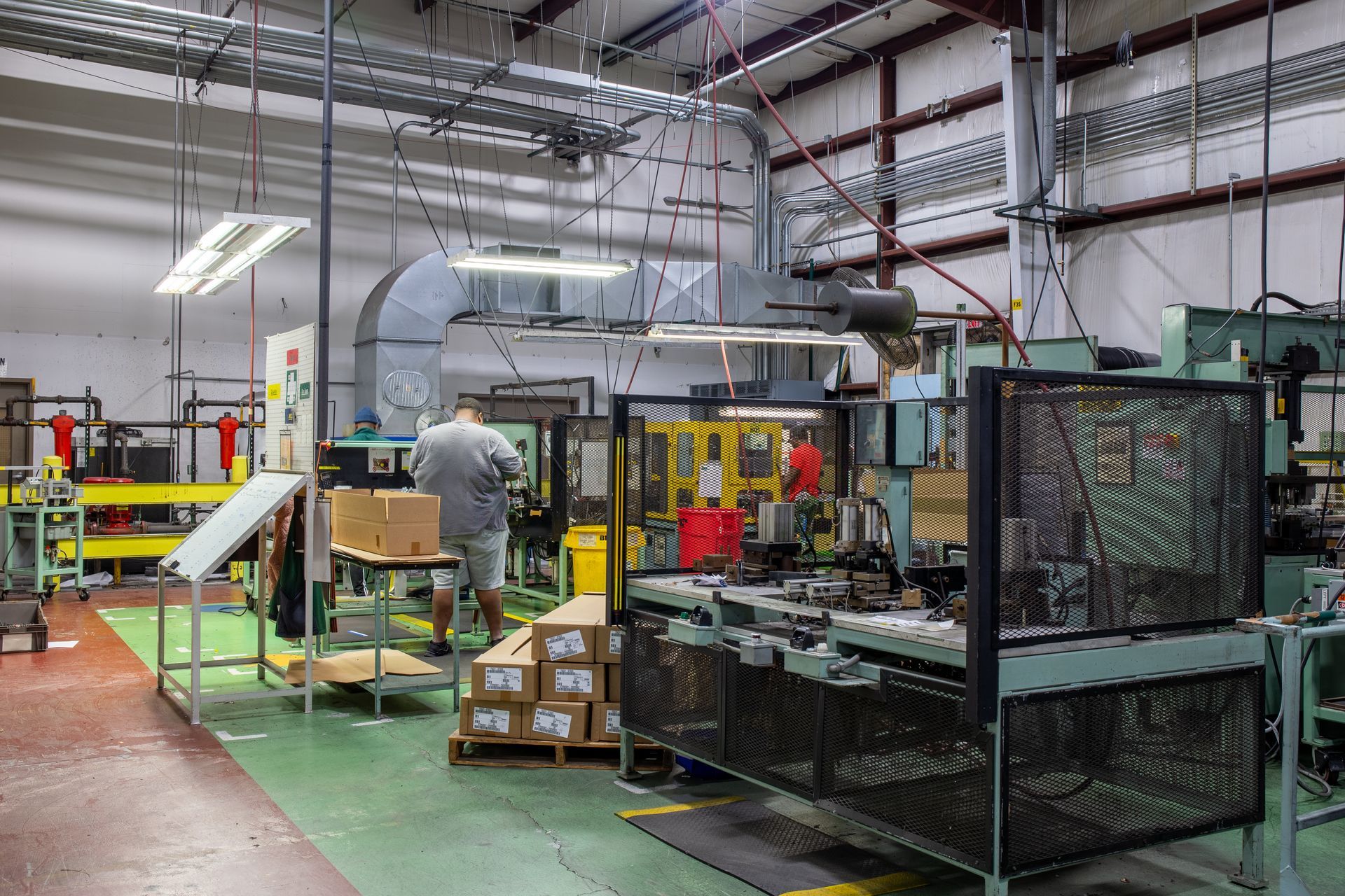 Factory interior with machinery, a worker, and boxes, with green and red floors, and overhead ductwork.