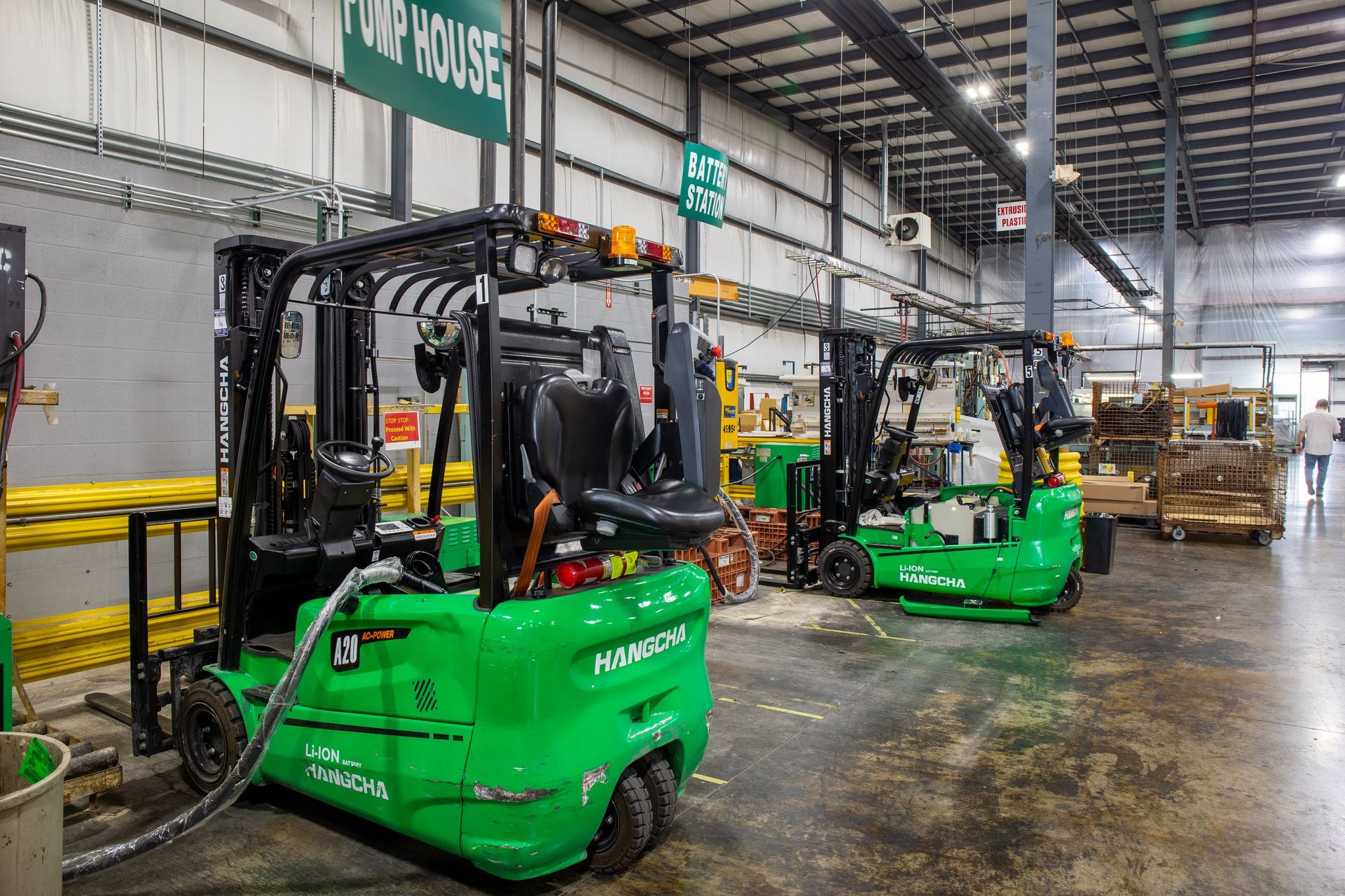 Two green forklifts inside a warehouse, a person walks near pallets.