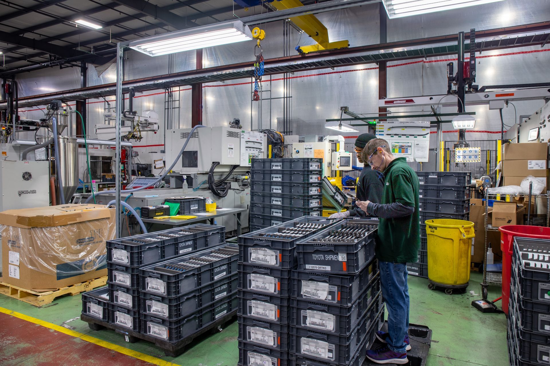 Factory interior: Two workers inspect black crates, machinery, and equipment on a production floor.