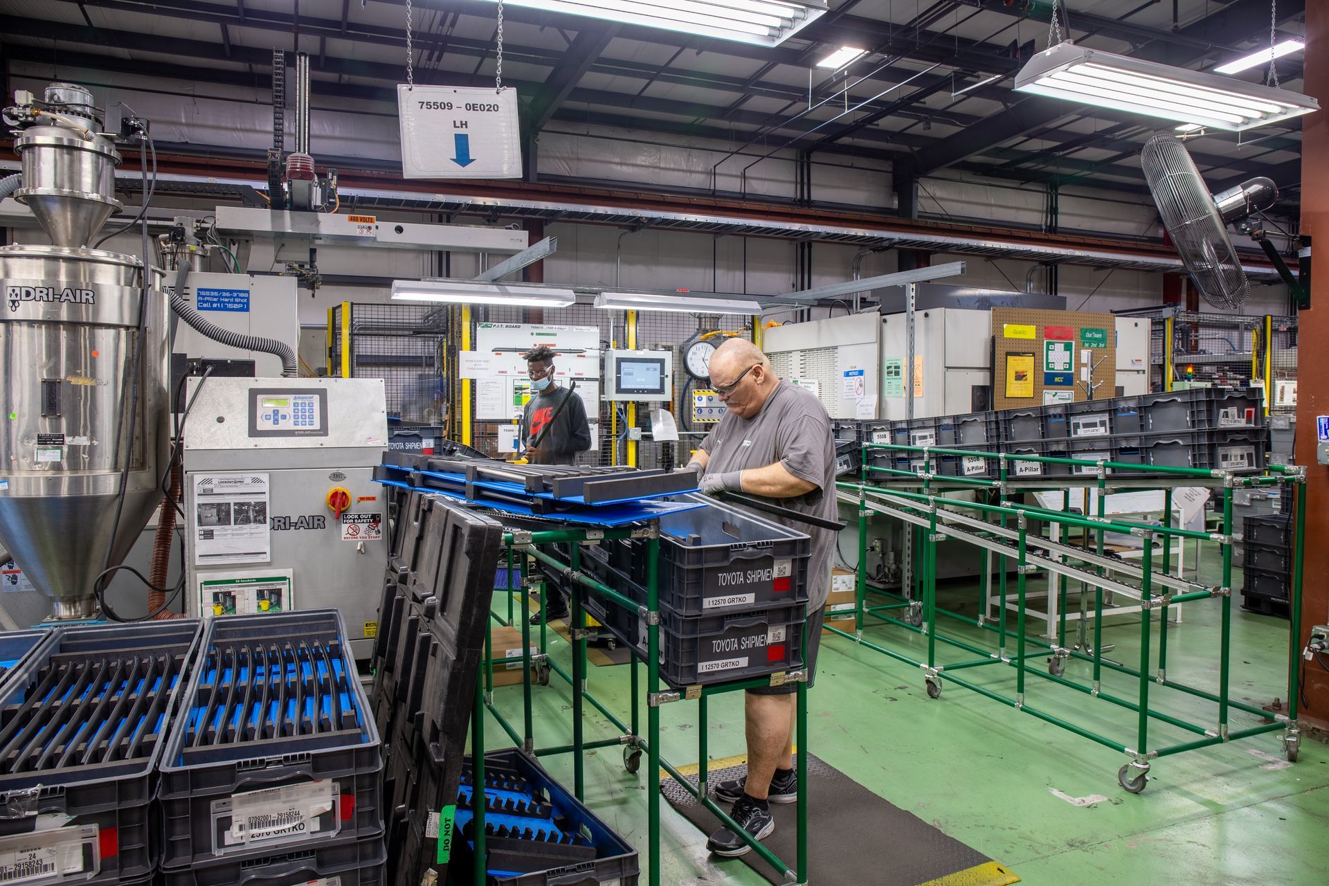 Factory interior: Workers assembling items at production line, with machines, conveyor belt, and stacks of bins.