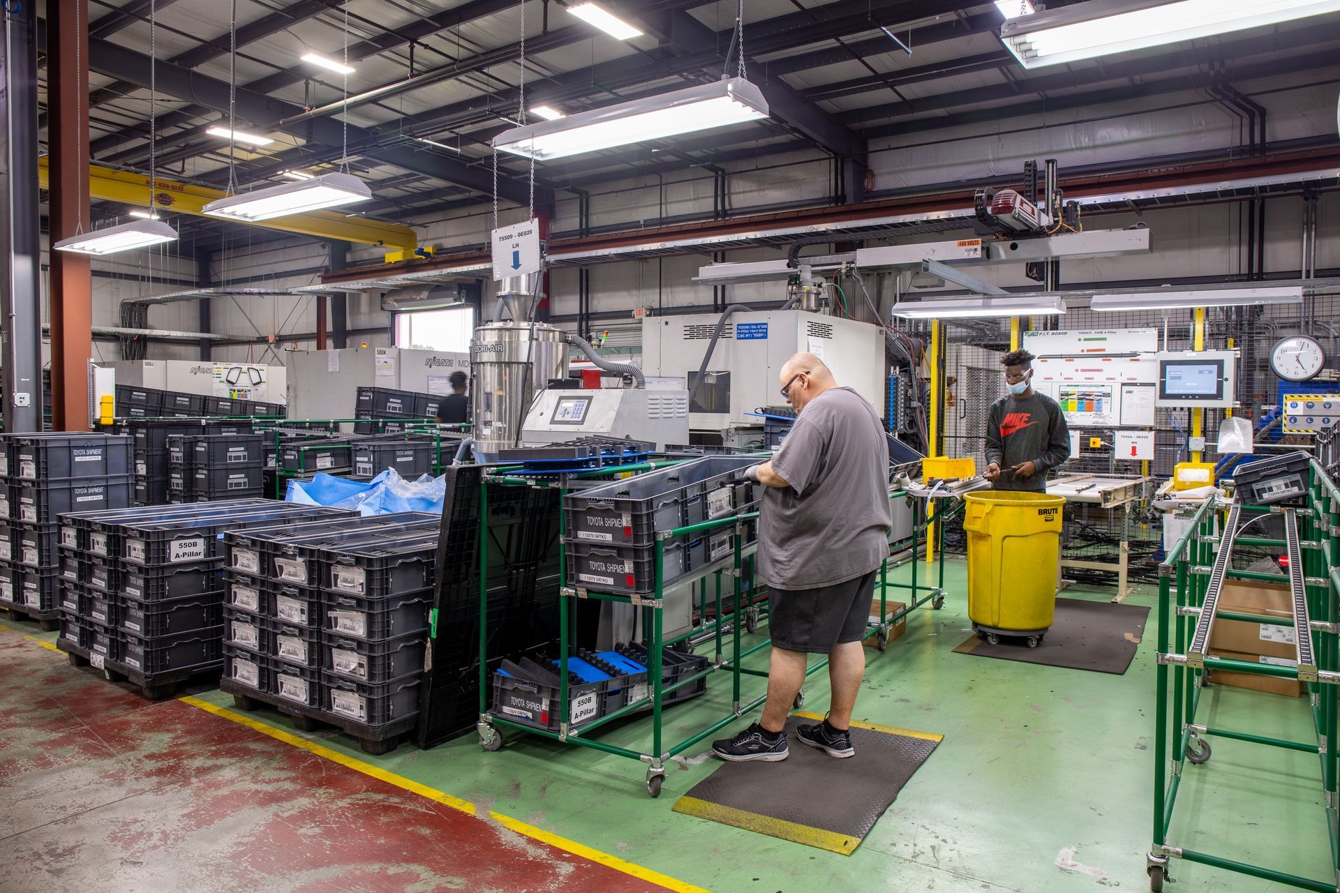 Factory interior: Workers near machinery and stacks of black bins.