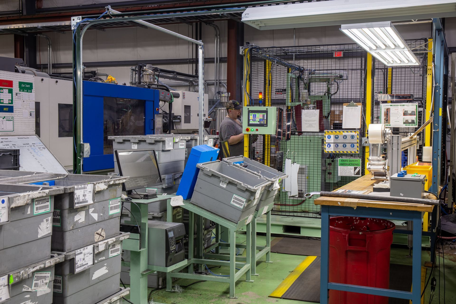 Factory floor with machinery, a worker, storage bins, and a red trash can.