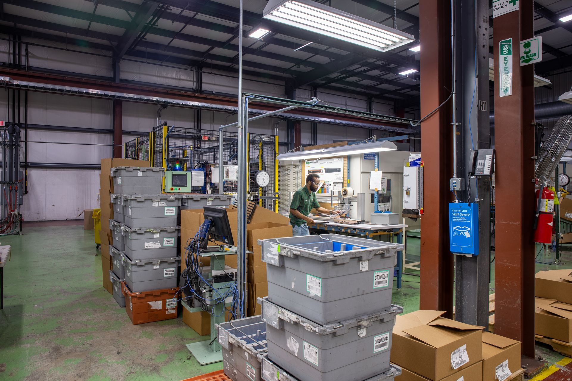 Warehouse interior with stacked gray bins, a man working at a workbench, and cardboard boxes.