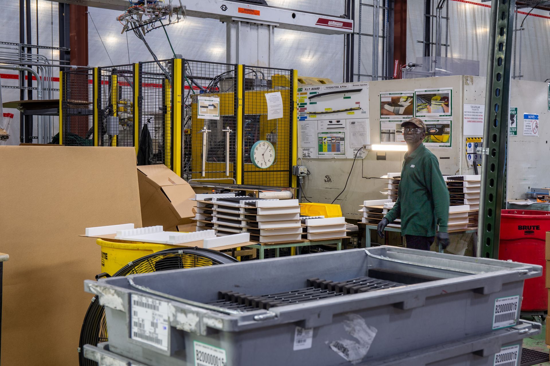 Factory worker in green shirt stands near machinery and bins filled with metal parts.