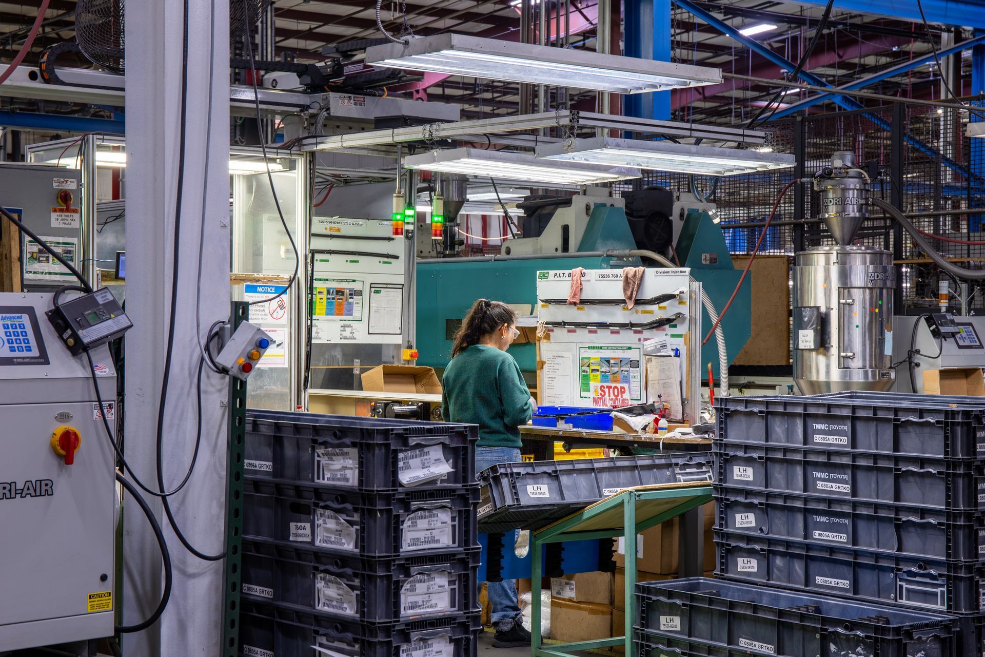 Factory worker standing at a workbench, surrounded by bins. Machinery and overhead lights in the background.