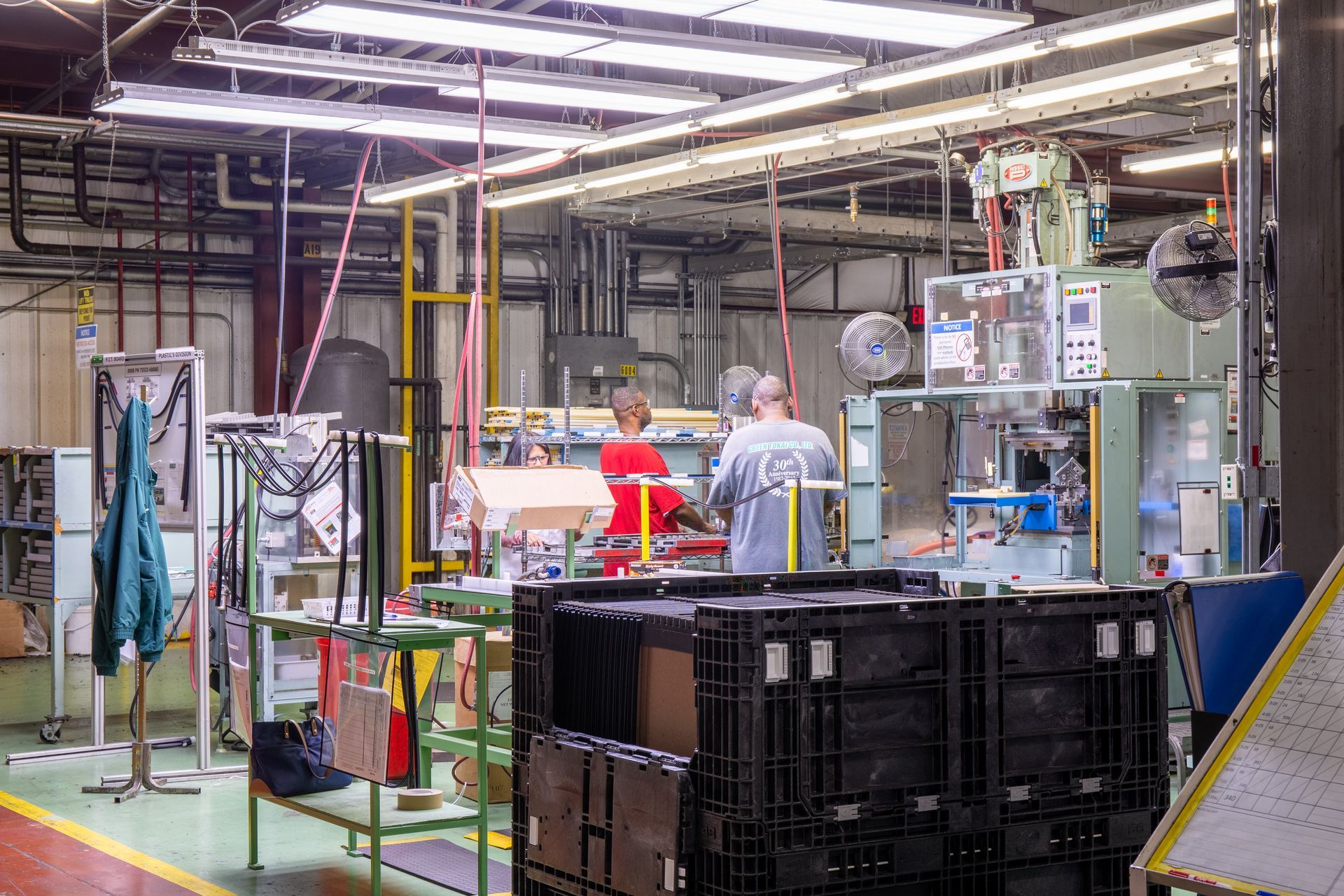 Factory interior with workers near machinery, plastic bins, and overhead lighting.