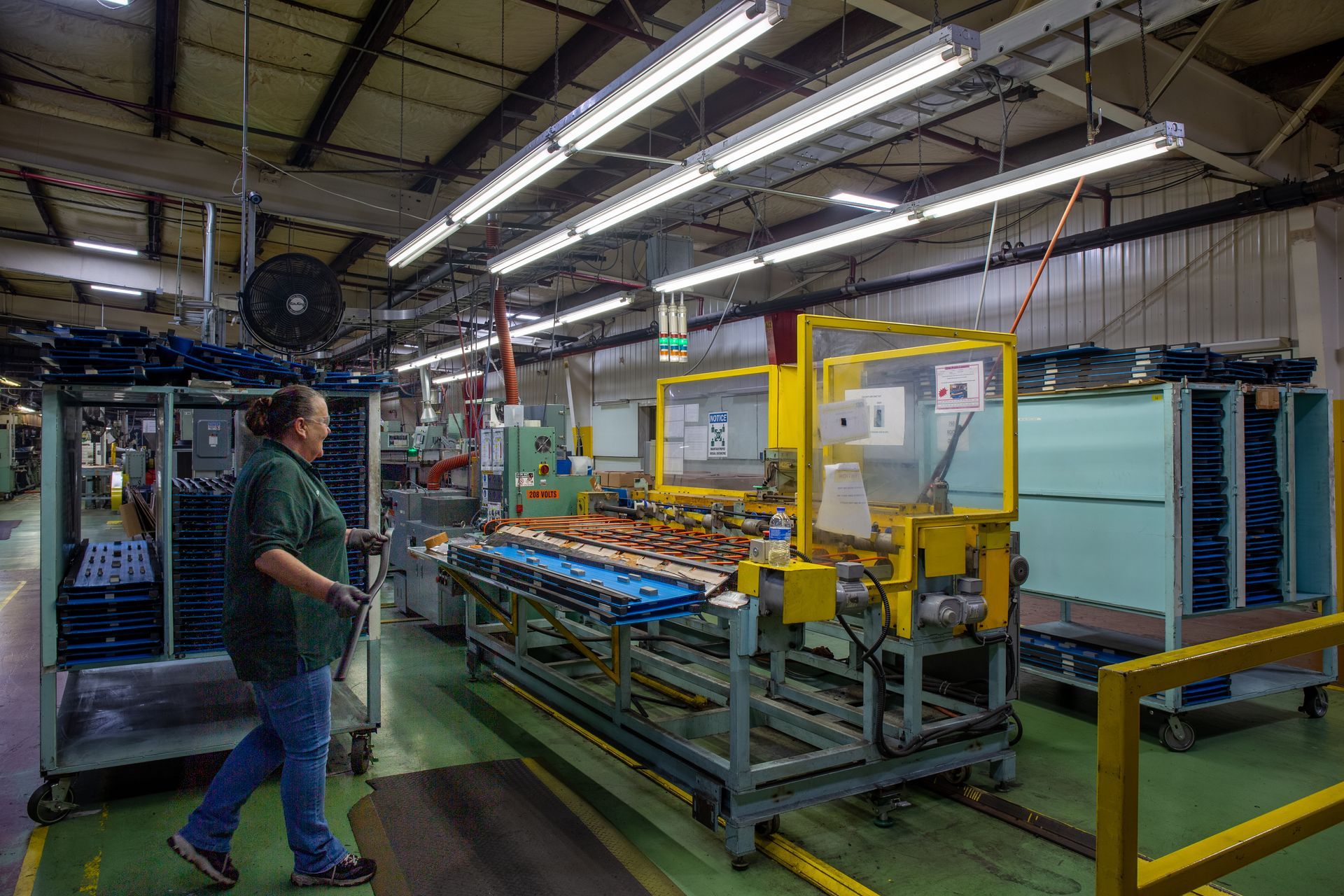 Factory worker operates machinery in a manufacturing facility, with product assembly lines and fluorescent lighting.
