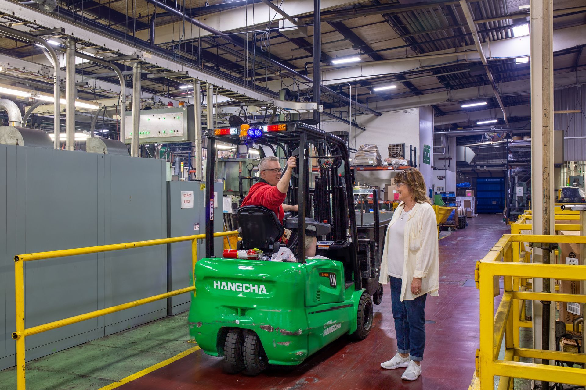 A person drives a green forklift in a factory, talking to a person standing nearby.
