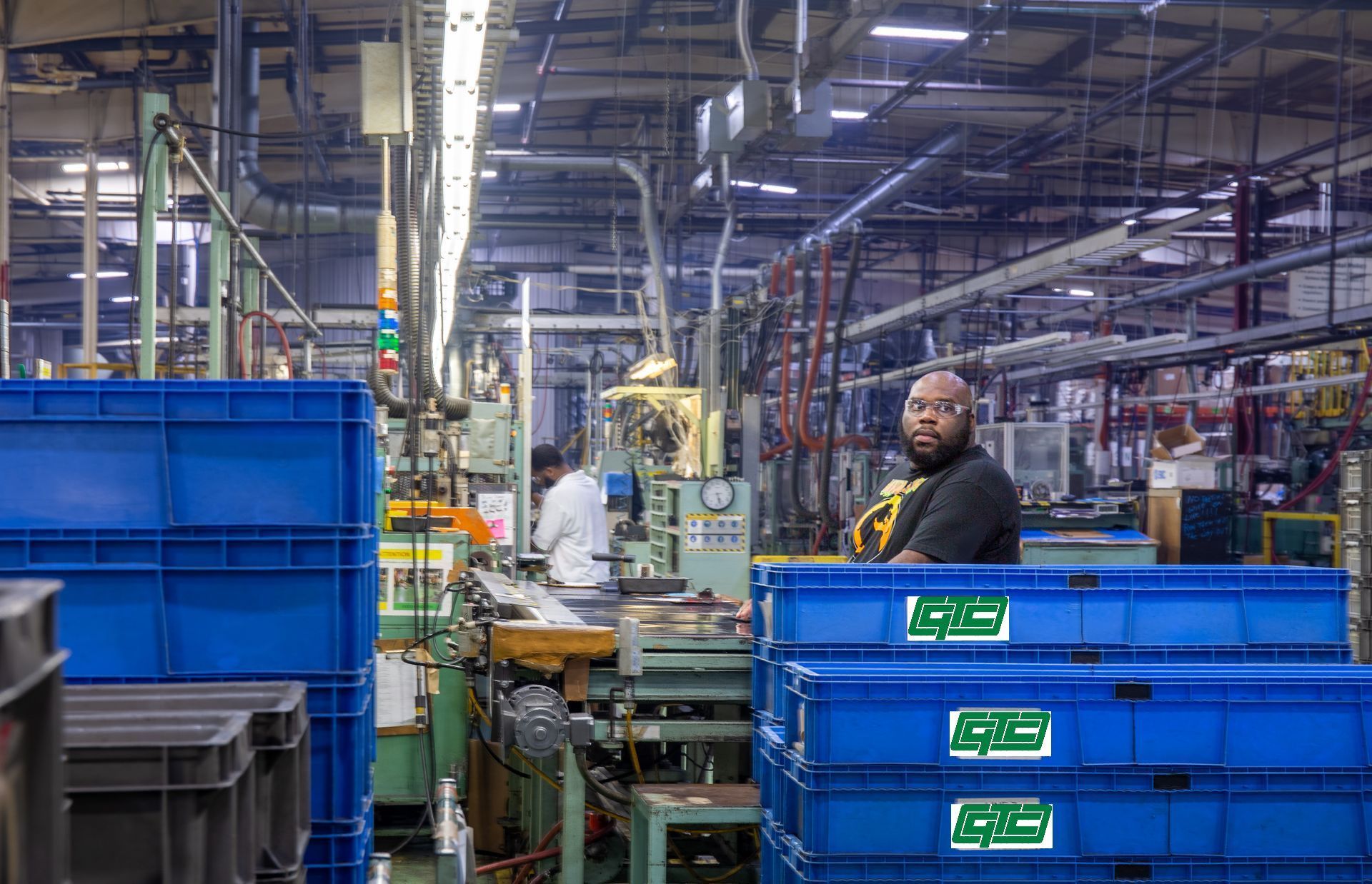 Man in black shirt at a factory, looking at the camera. Blue bins and machinery fill the interior.