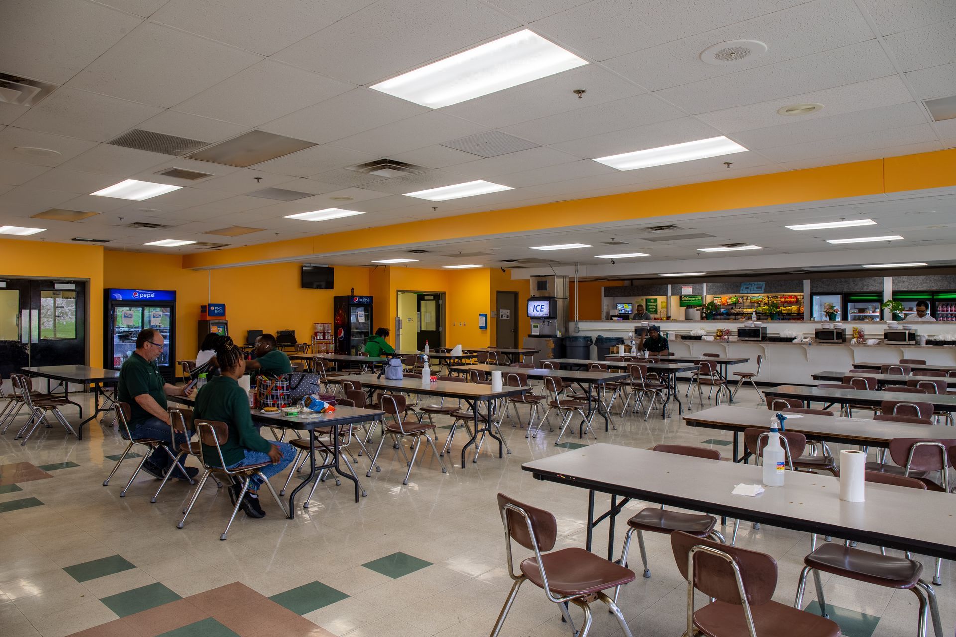 Dining hall with tables and chairs, bright lighting, and a few people seated.
