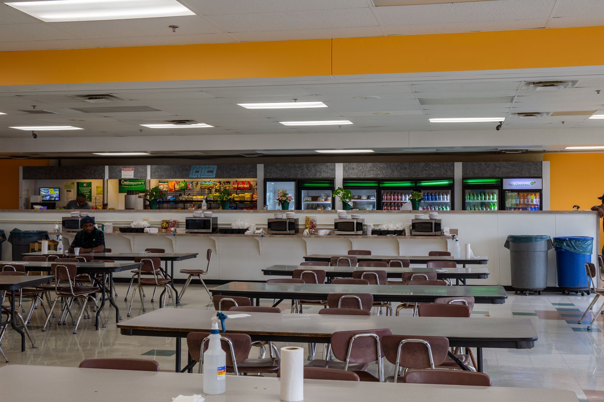 Empty cafeteria with tables, chairs, and a serving counter with coolers and food.