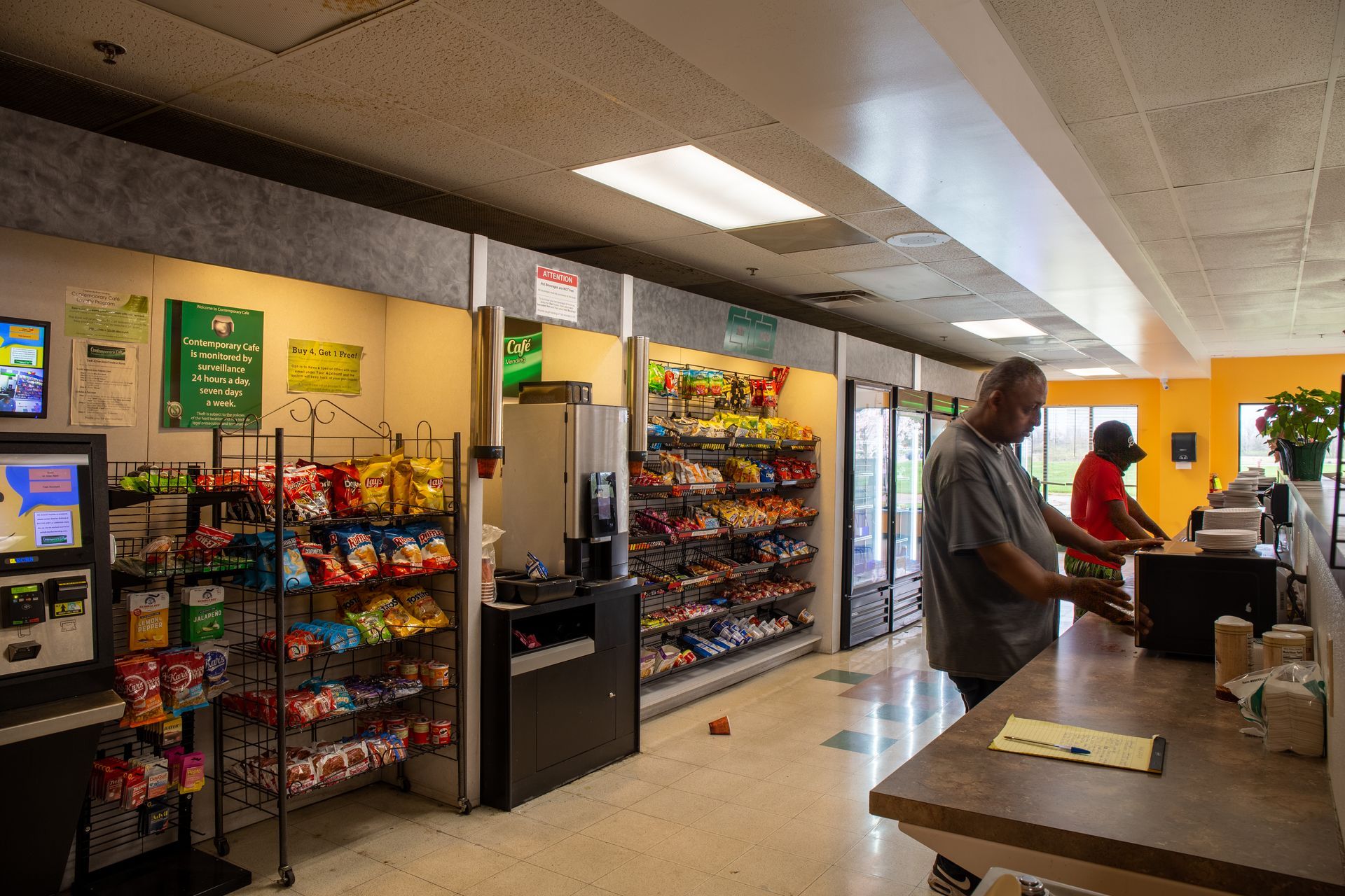 Inside a convenience store. Person paying at the counter while another person browses snacks.