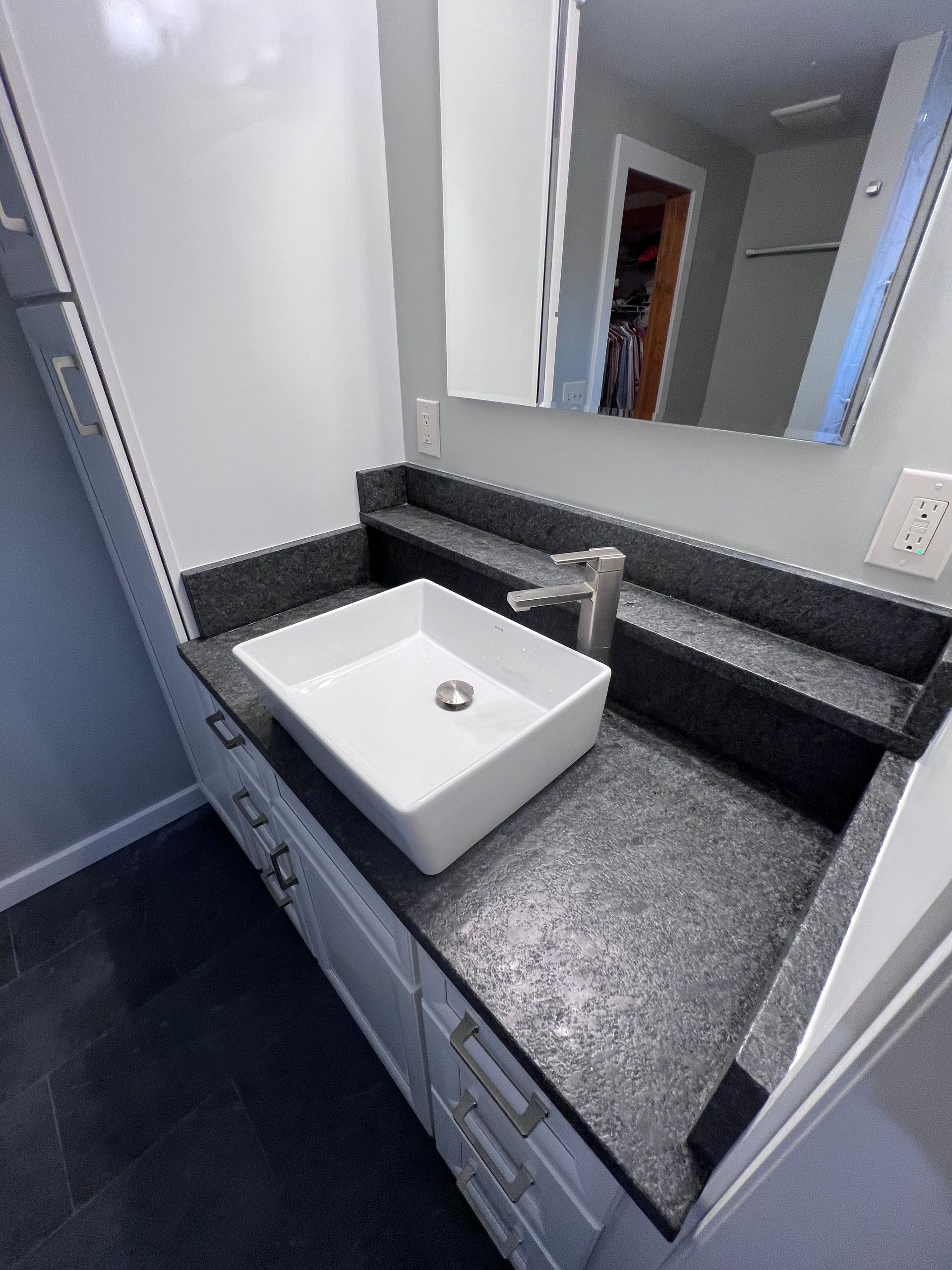 Modern bathroom vanity with white sink, dark granite countertop, mirror, and light gray cabinetry