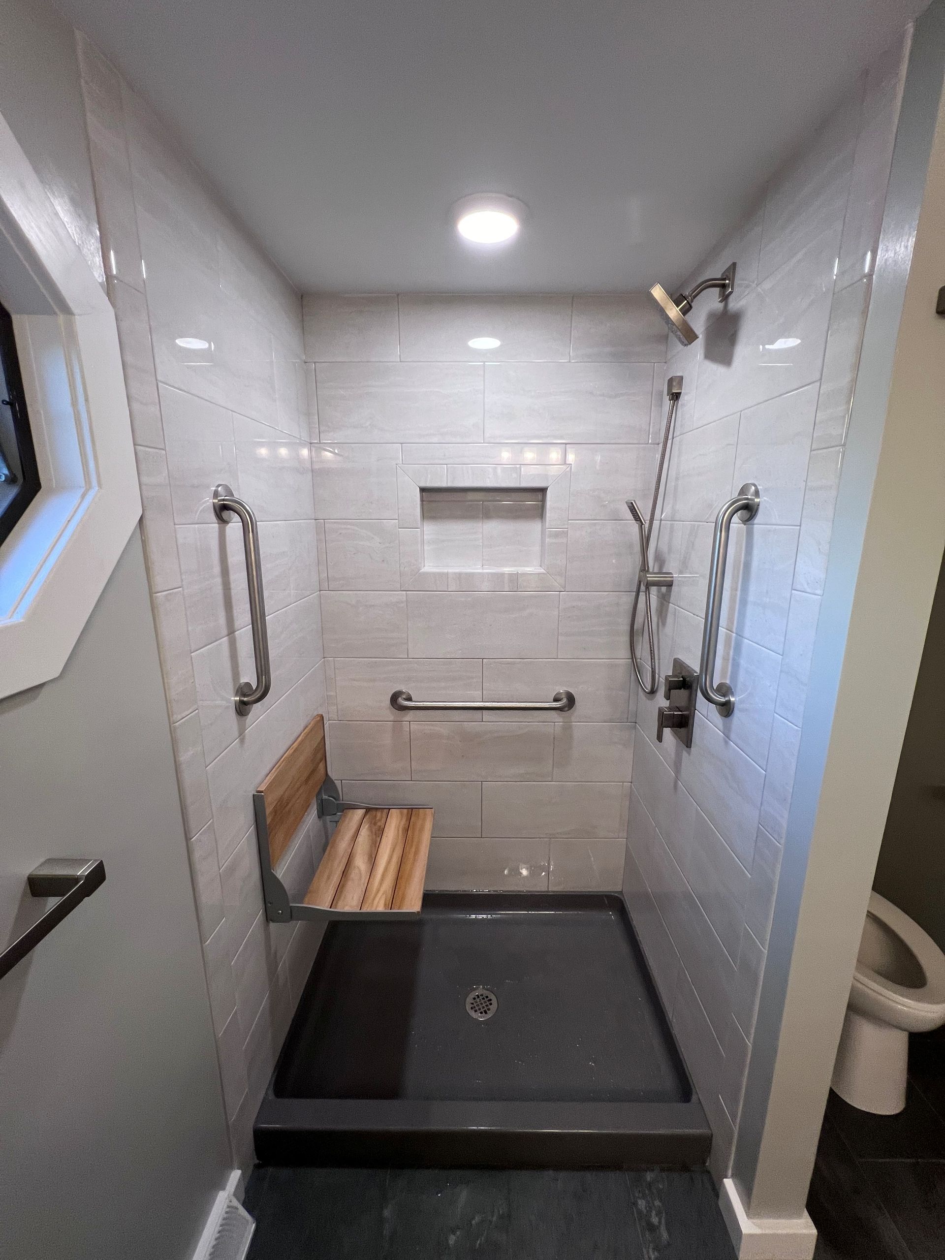 Accessible shower stall with white tile walls, grab bars, built-in bench, and a handheld showerhead.