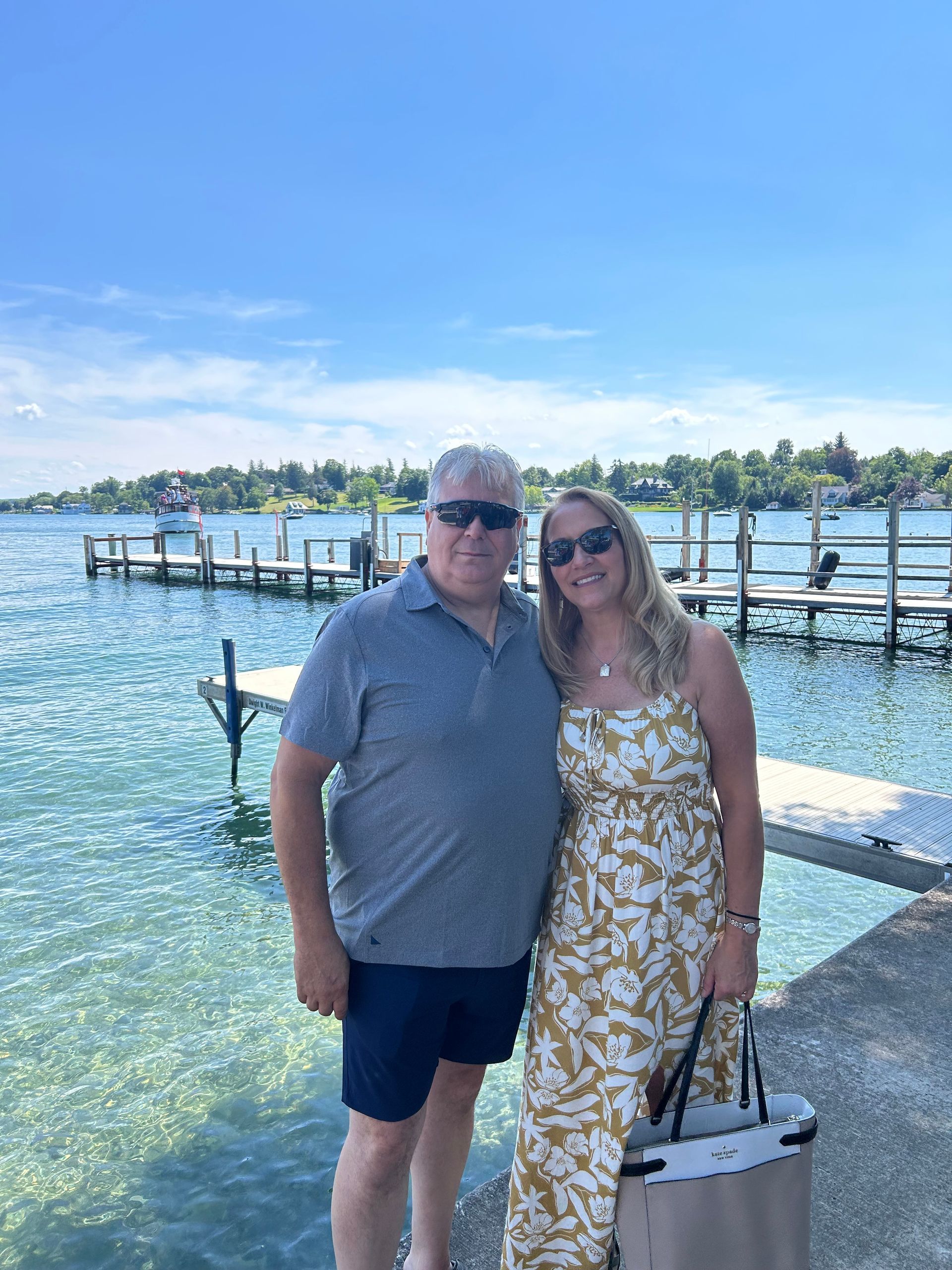 Two people standing by a lakeside dock, smiling, with blue water and a bright sky behind them.