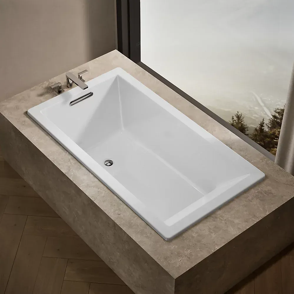 Modern white bathtub set into beige stone platform beside a window with a forest view