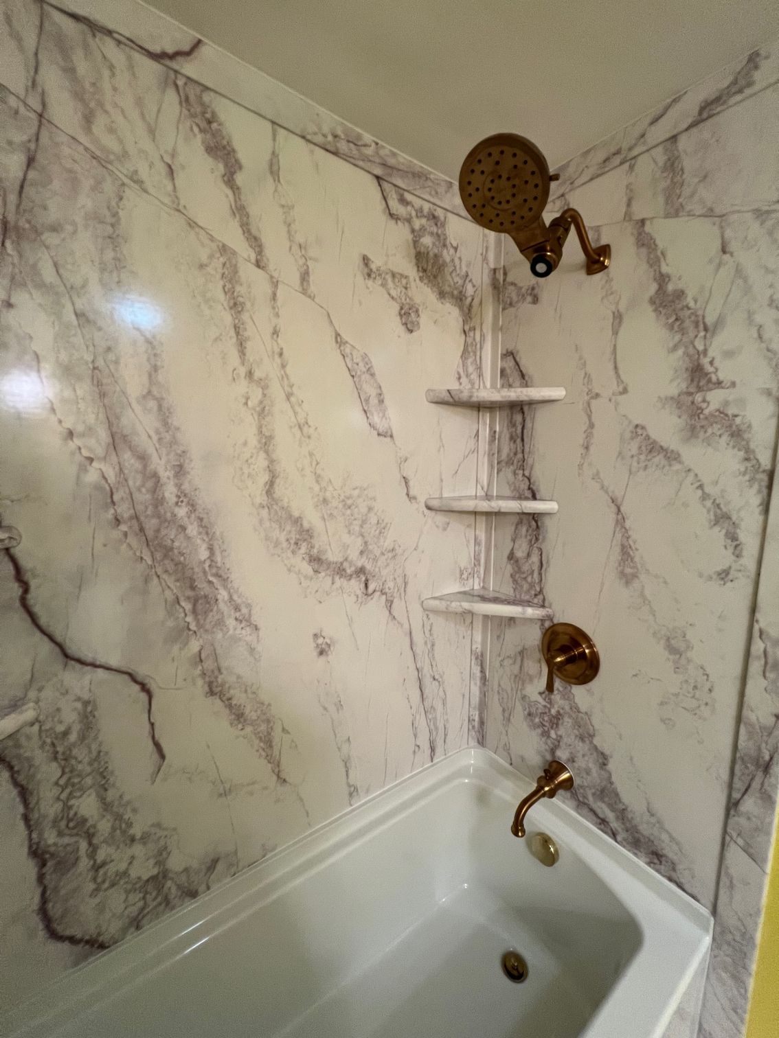 Corner shower with marble-look walls, built-in shelves, and a bathtub below a showerhead.