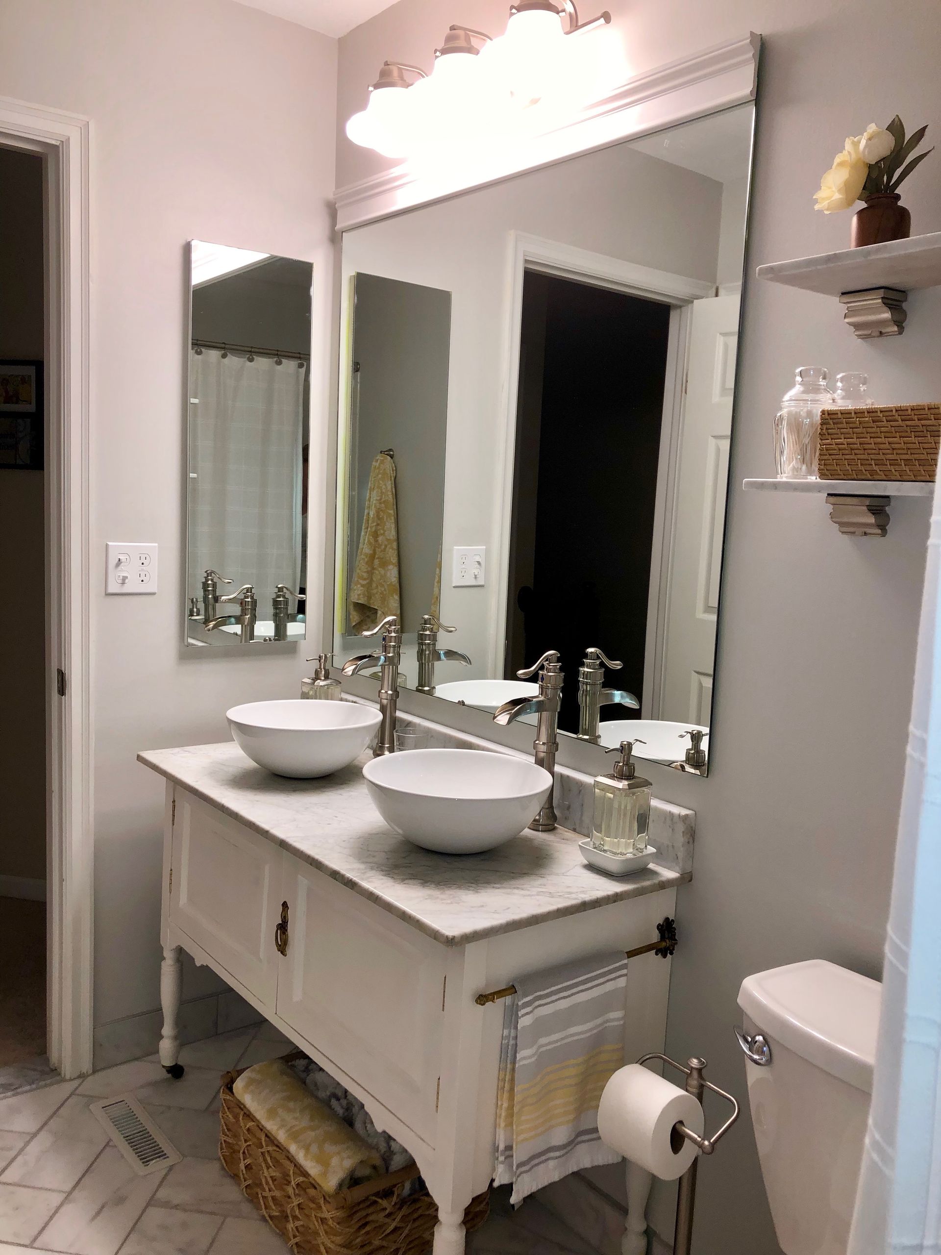 Bright bathroom vanity with twin sinks, large mirror, shelves, and white tile flooring