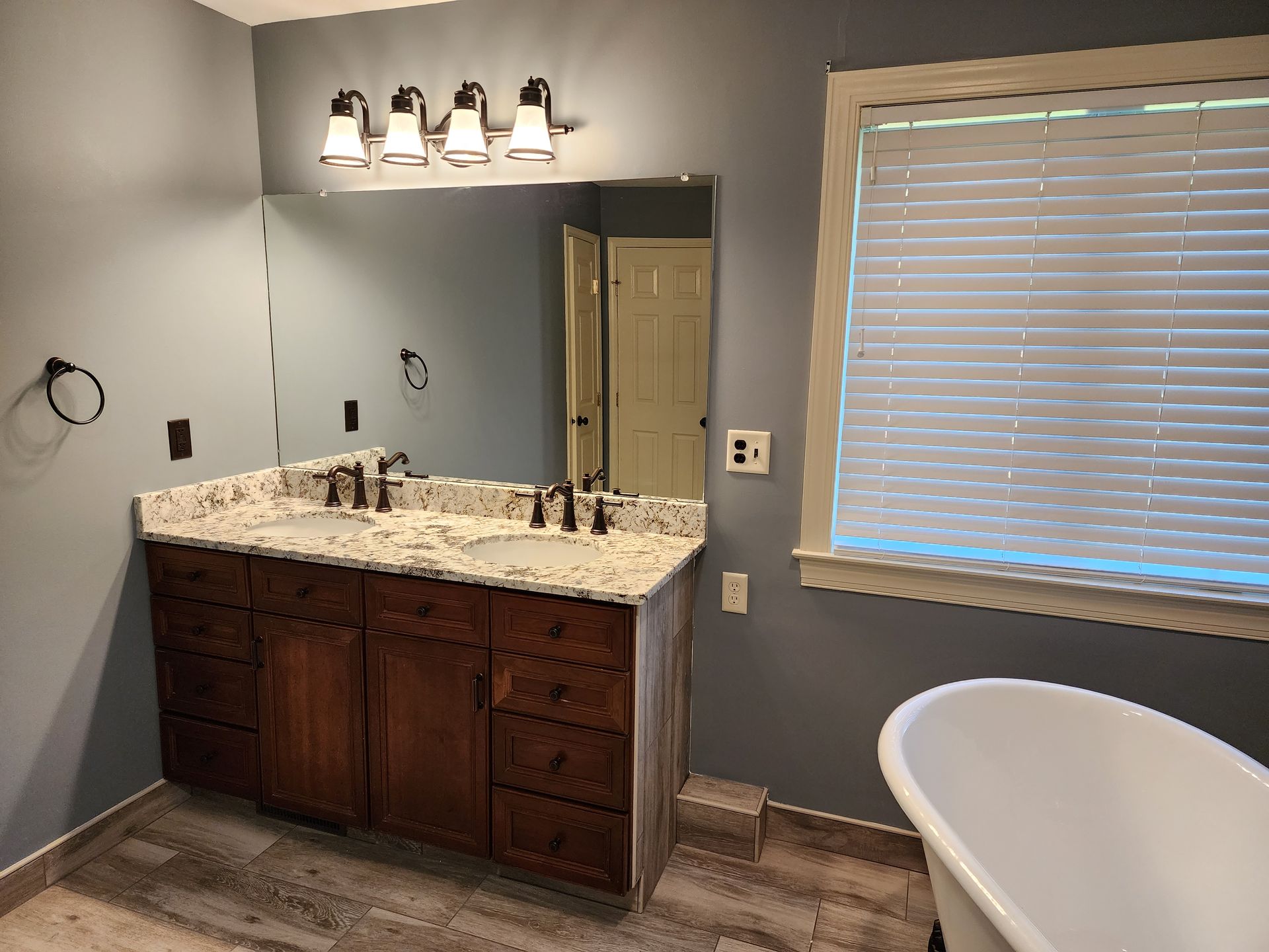 Bathroom with granite vanity, dark wood cabinets, wall mirror, light fixture, and window blinds
