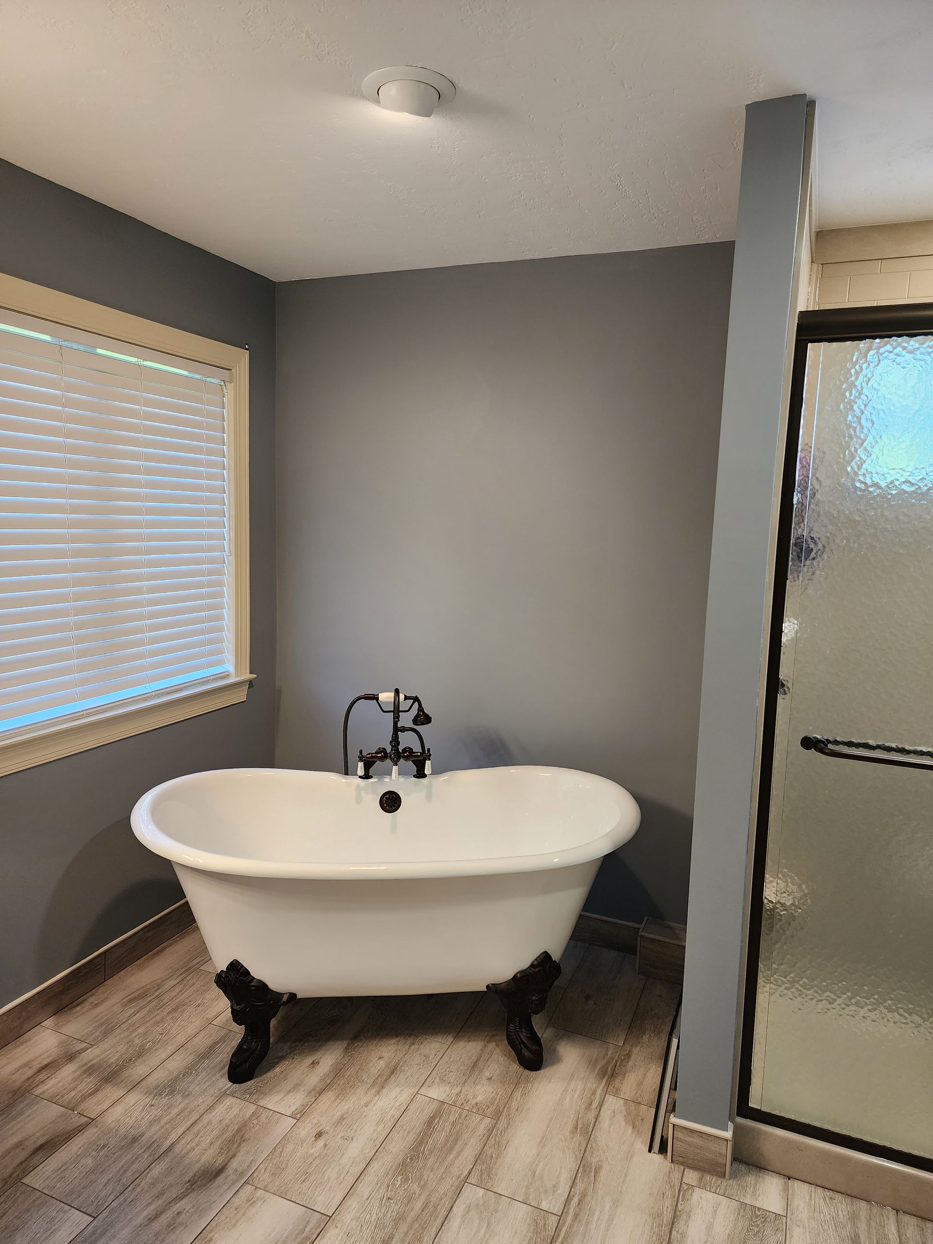 Clawfoot bathtub in a gray bathroom beside a frosted glass shower and window blinds