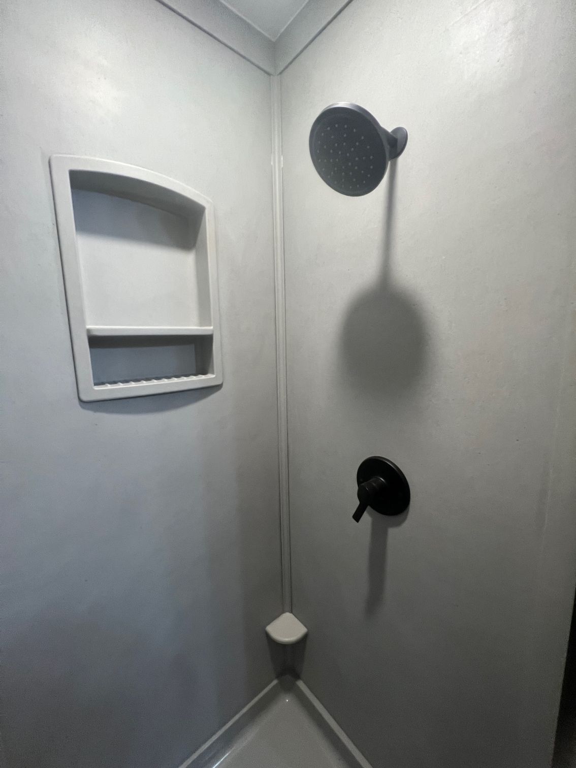 Small gray shower stall with a wall shelf, showerhead, control knob, and corner shelf.