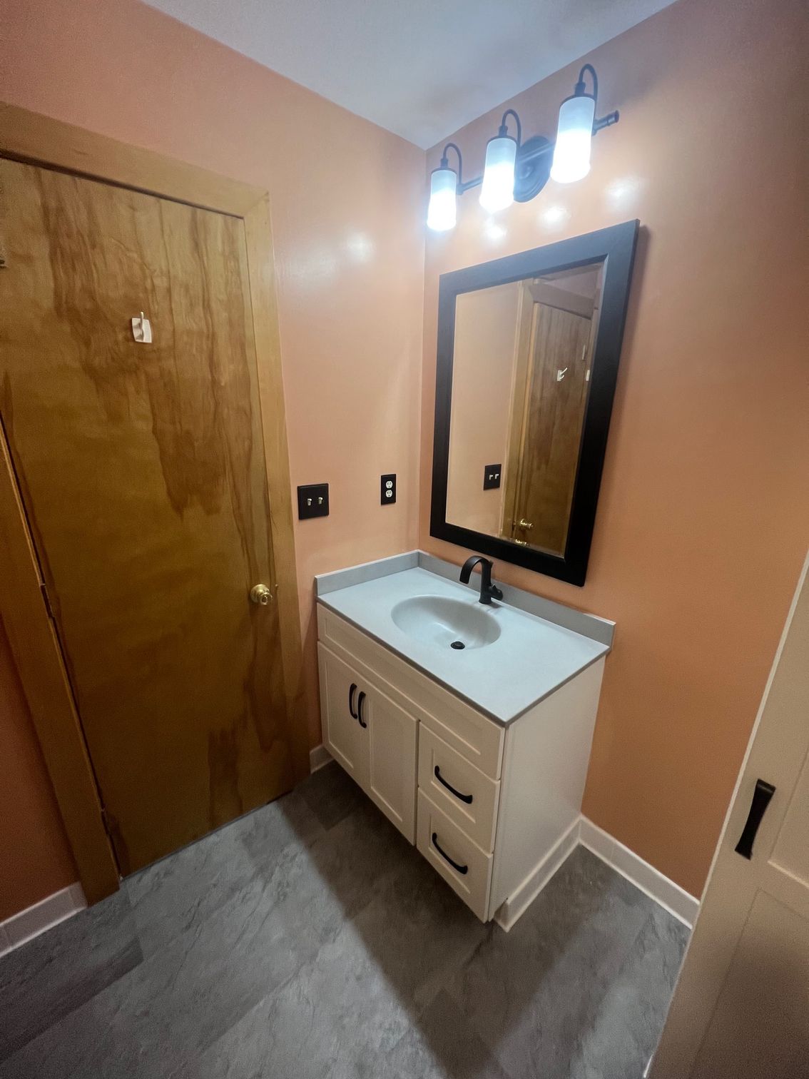 Bathroom vanity with sink and mirror beside a wooden door, in a peach-colored room.