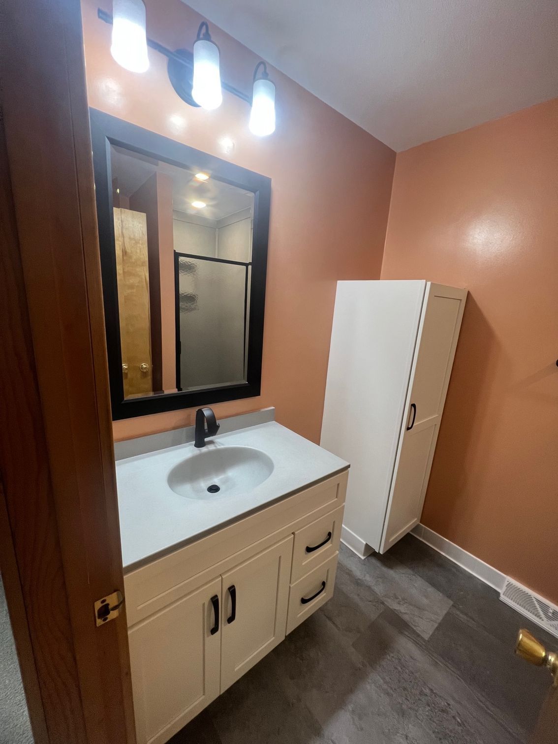 Small bathroom vanity with mirror, white sink, peach walls, and a tall white cabinet on the right