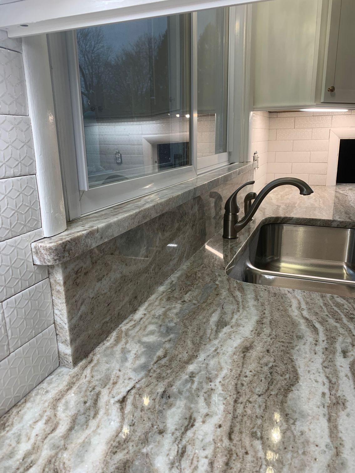 Kitchen countertop and sink with gray marble pattern, white tile backsplash, and a window above the counter