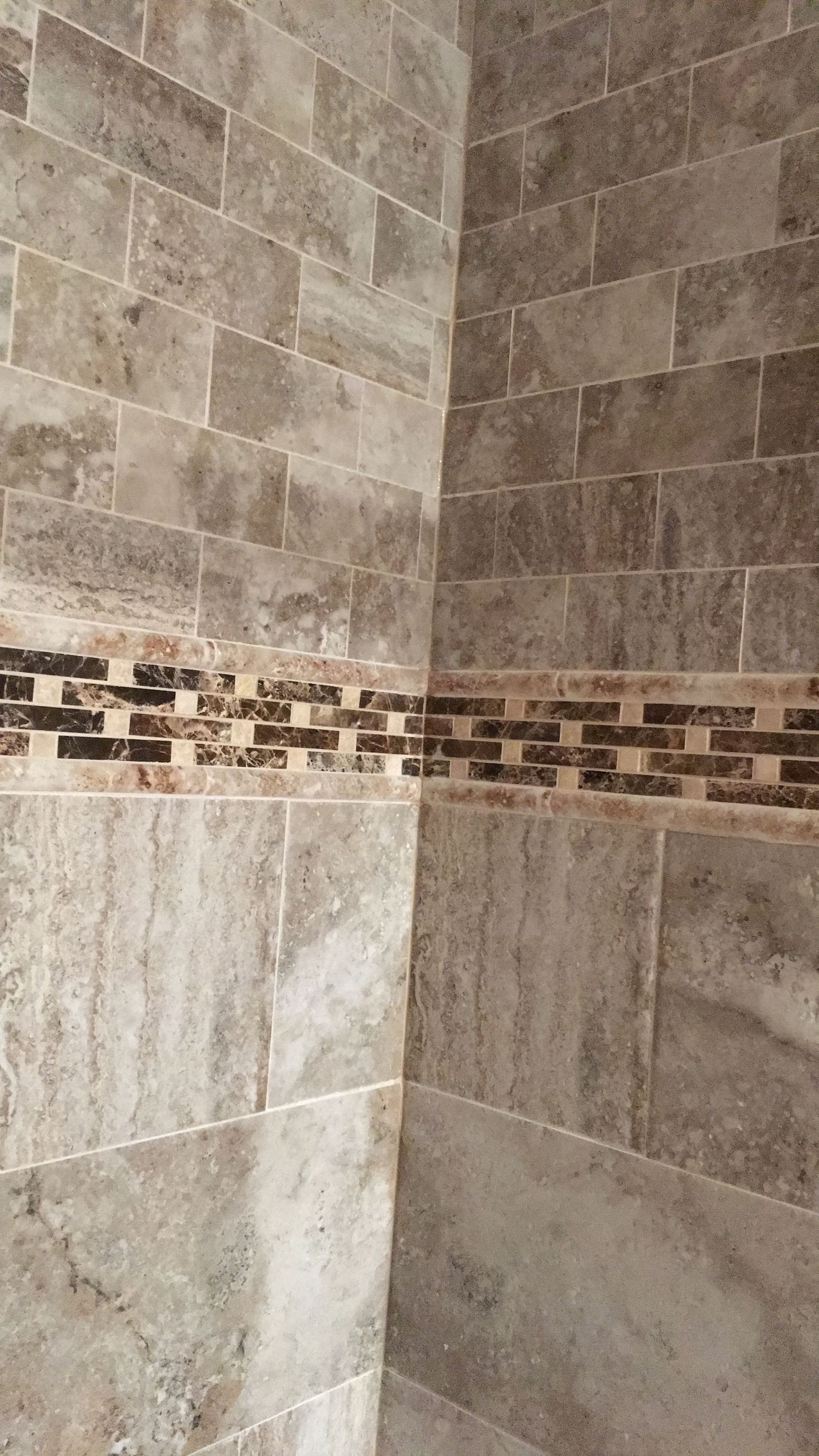Corner of a tiled shower wall with beige stone tiles and a decorative brown mosaic border