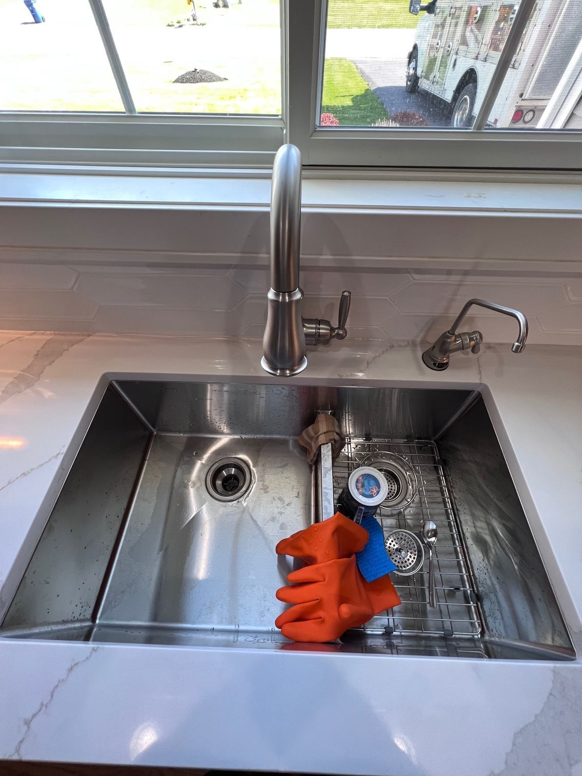 Kitchen sink with orange gloves, dish rack, and sunlight by the window