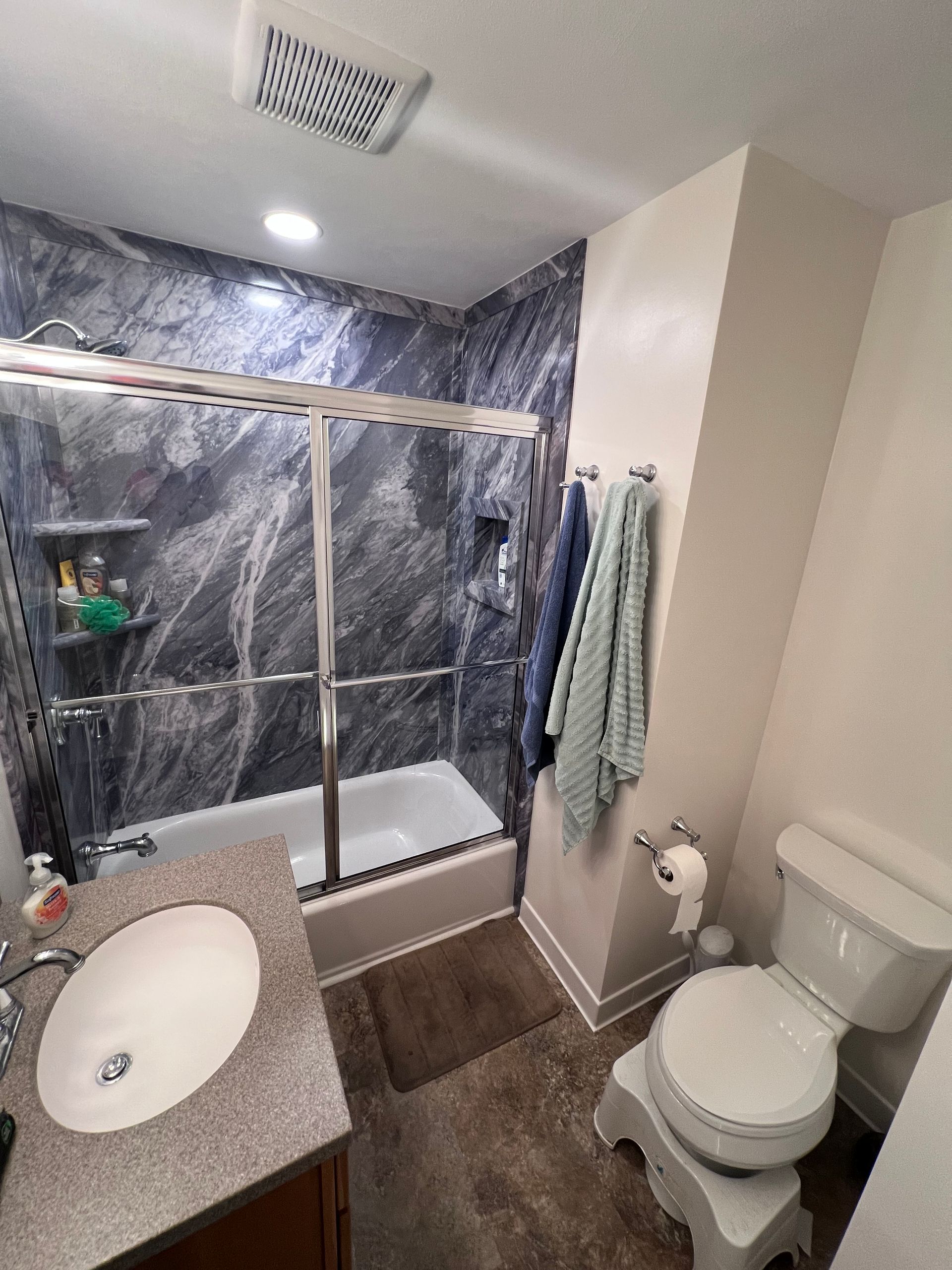 Small bathroom with marble-look shower, toilet, sink, and towels hanging on the wall.