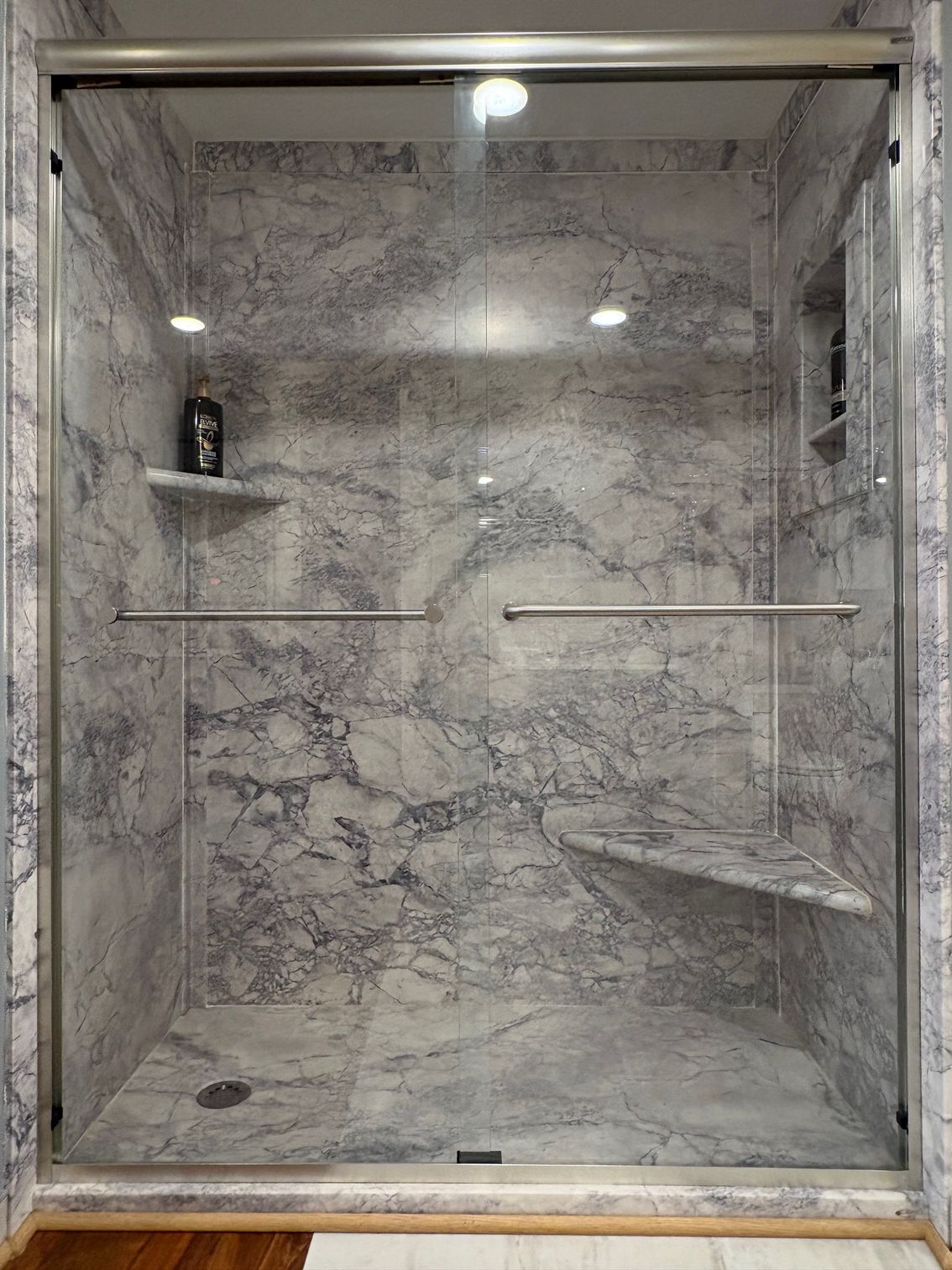 Modern marble-tiled walk-in shower with glass enclosure, shelves, and grab bars